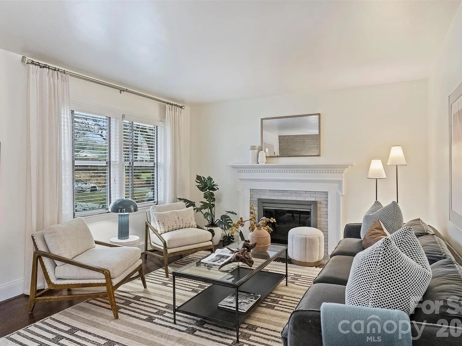 Bright living room with white walls, wood floors, gray sofa with patterned pillows, two beige armchairs, glass coffee table, white fireplace with mirror, and large window with white curtains.