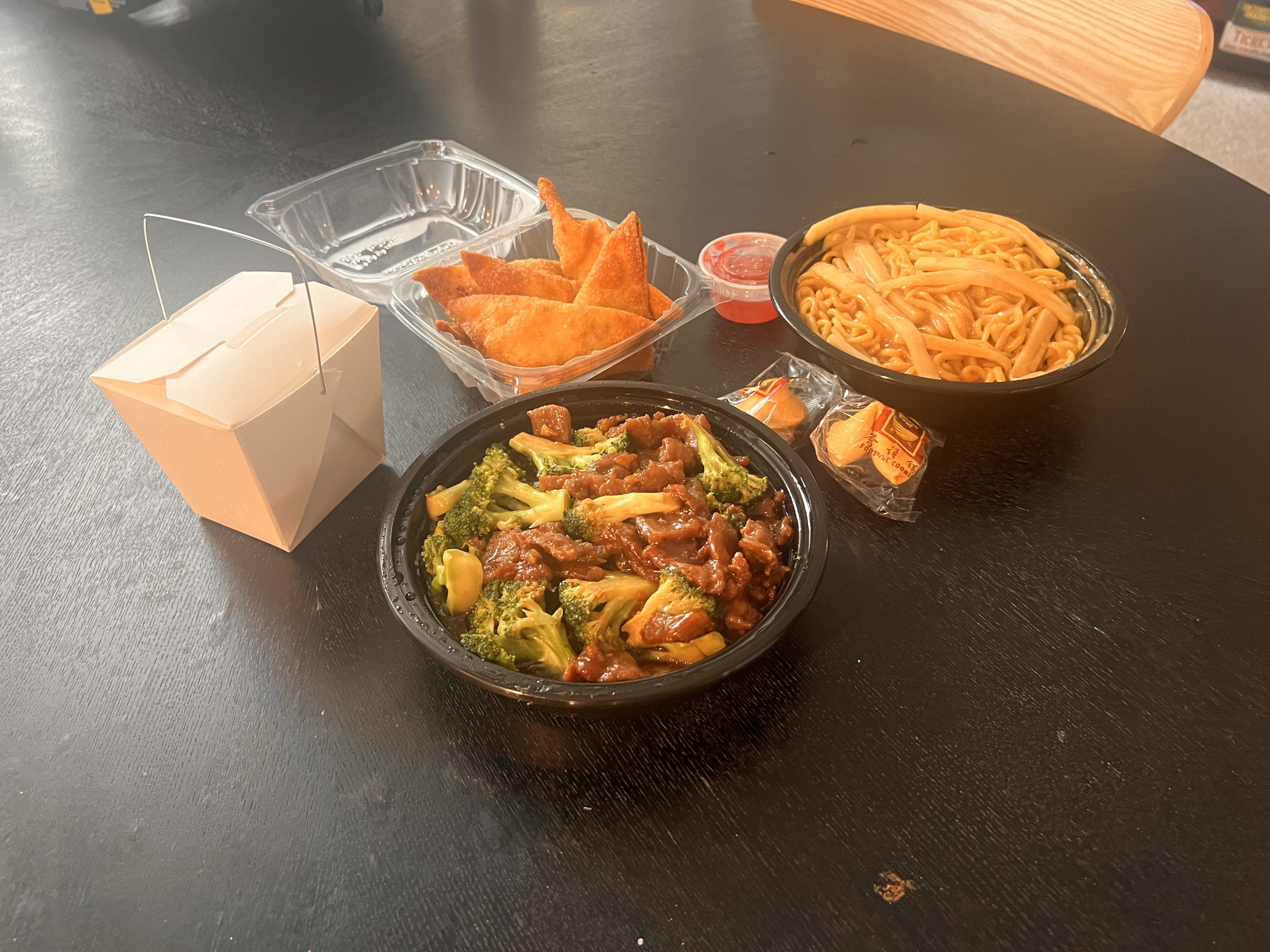 Chinese takeout meal with beef and broccoli in a black bowl, crispy wontons in a plastic container with red sauce, noodles with bamboo shoots, a white takeout box, and two fortune cookies on a black table.