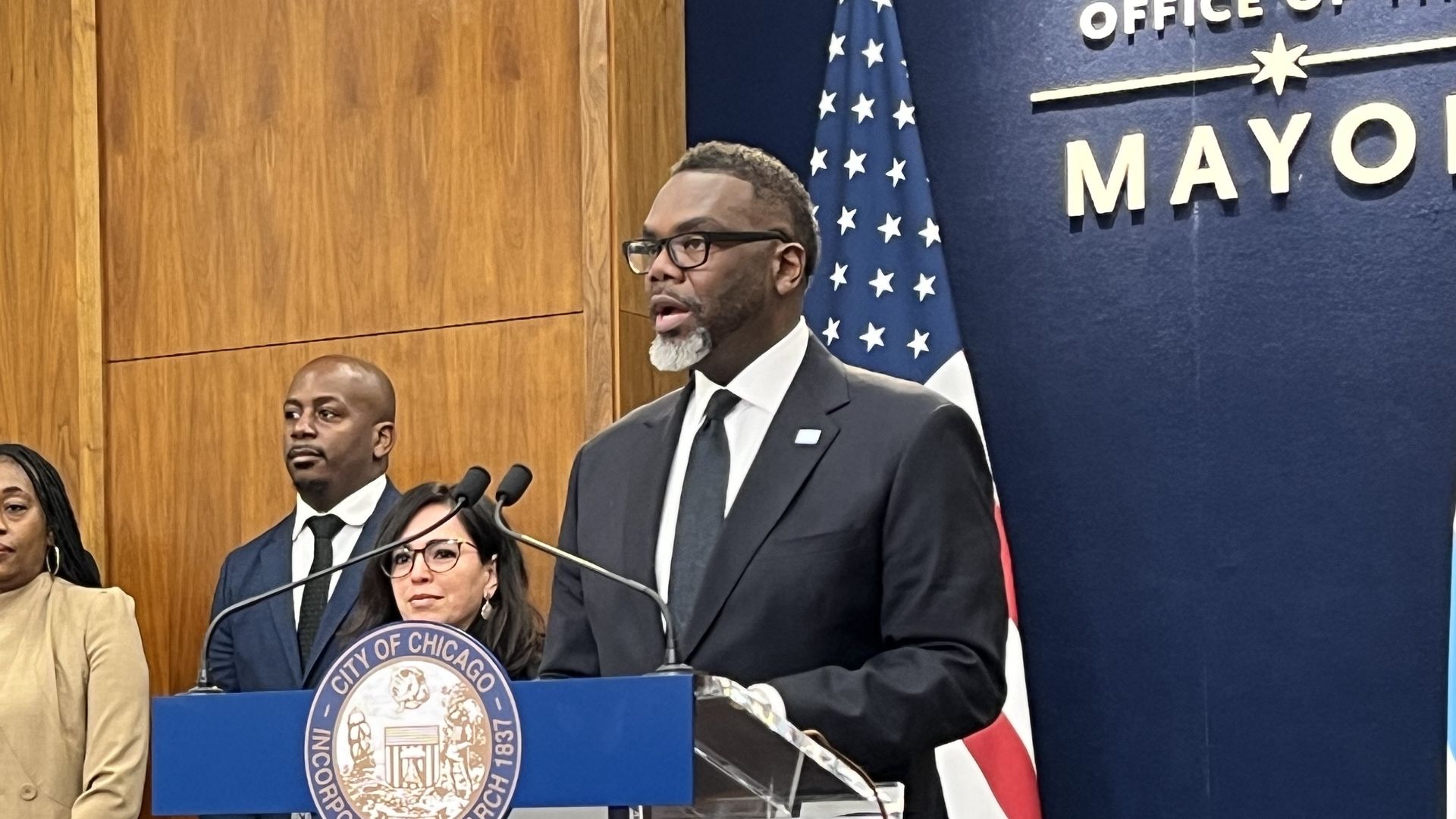 A man in a dark suit and glasses speaks at a podium bearing the City of Chicago seal; officials stand nearby. An American flag and a blue backdrop reading "CITY OF CHICAGO" are behind him.