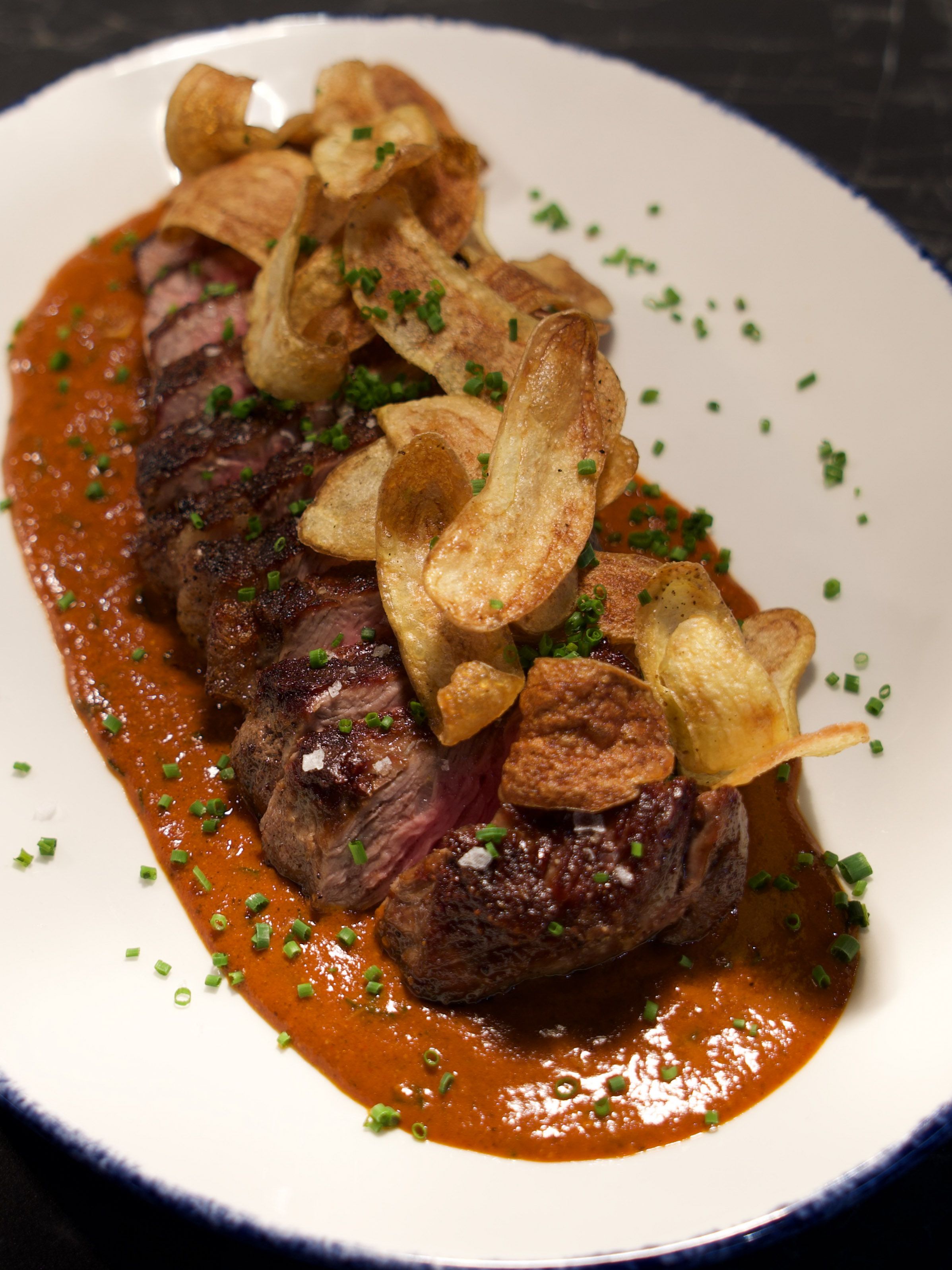Sliced grilled steak served on red sauce, topped with crispy potato chips and sprinkled with chopped green herbs on a white plate with a blue rim.