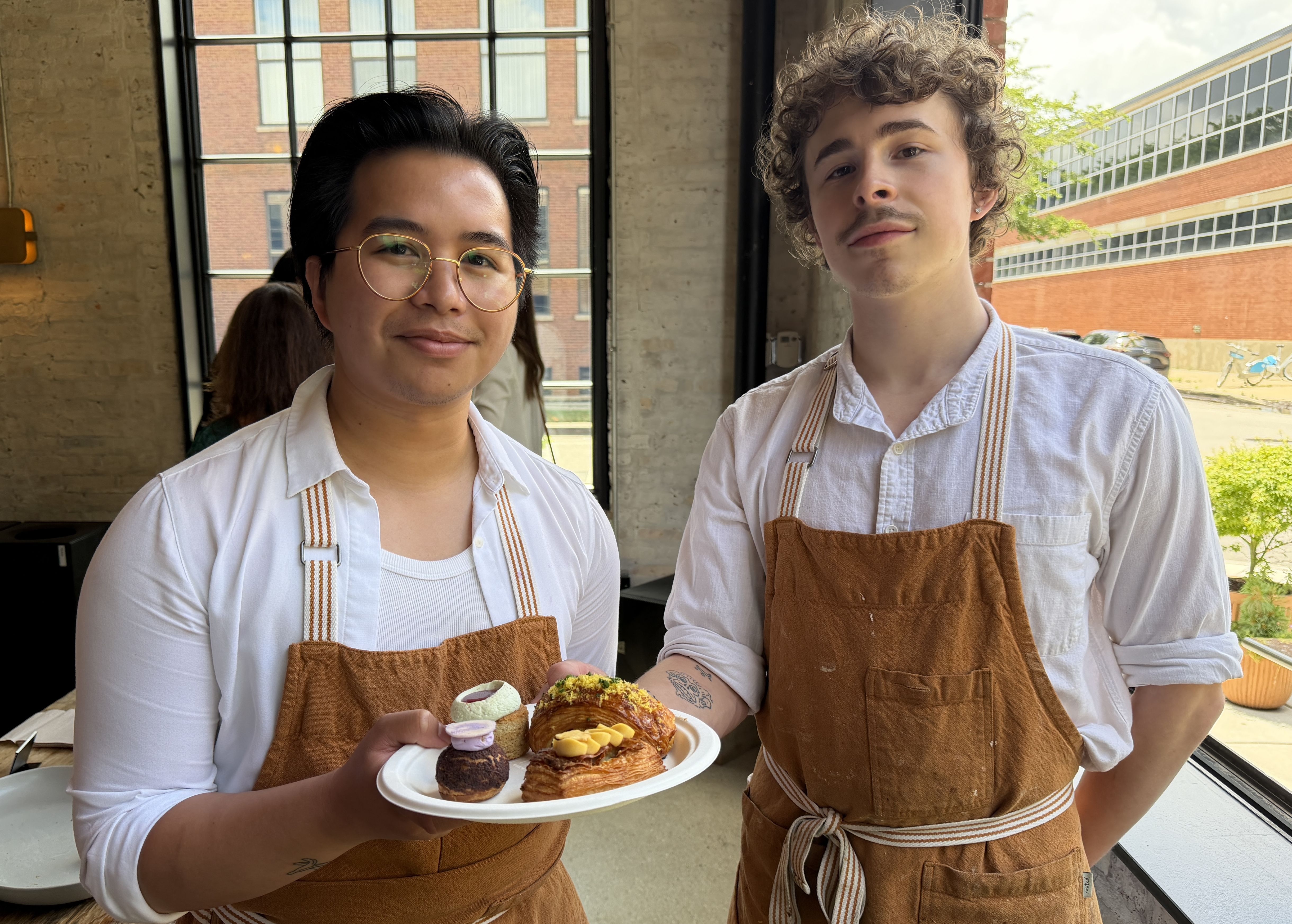 Two bakery workers in white shirts with brown aprons stand by a large window, one wearing round glasses and dark hair, the other with curly hair, presenting a plate of assorted pastries and macarons.