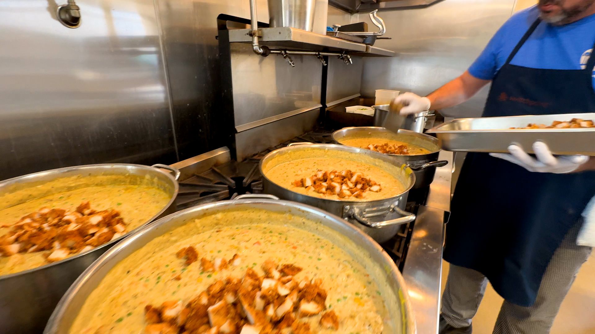 Four large pots on a stove filled with creamy yellow soup with vegetables, topped with pieces of cooked brown meat; a person wearing gloves, a blue shirt and black apron adds meat from a tray.
