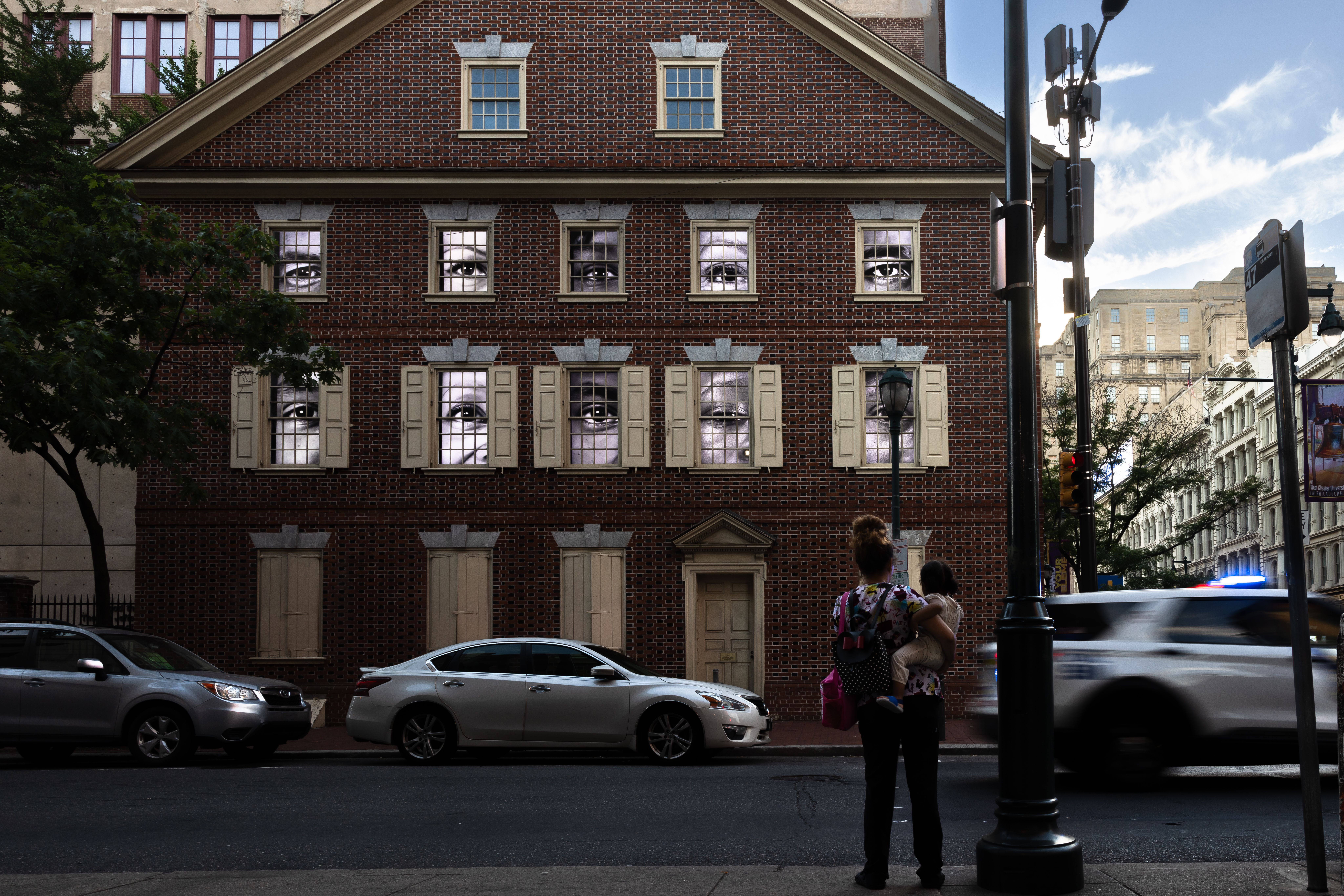 A woman holding a child stands across the street from the Declaration House, where black-and-white moving images of eyes fill the windows.