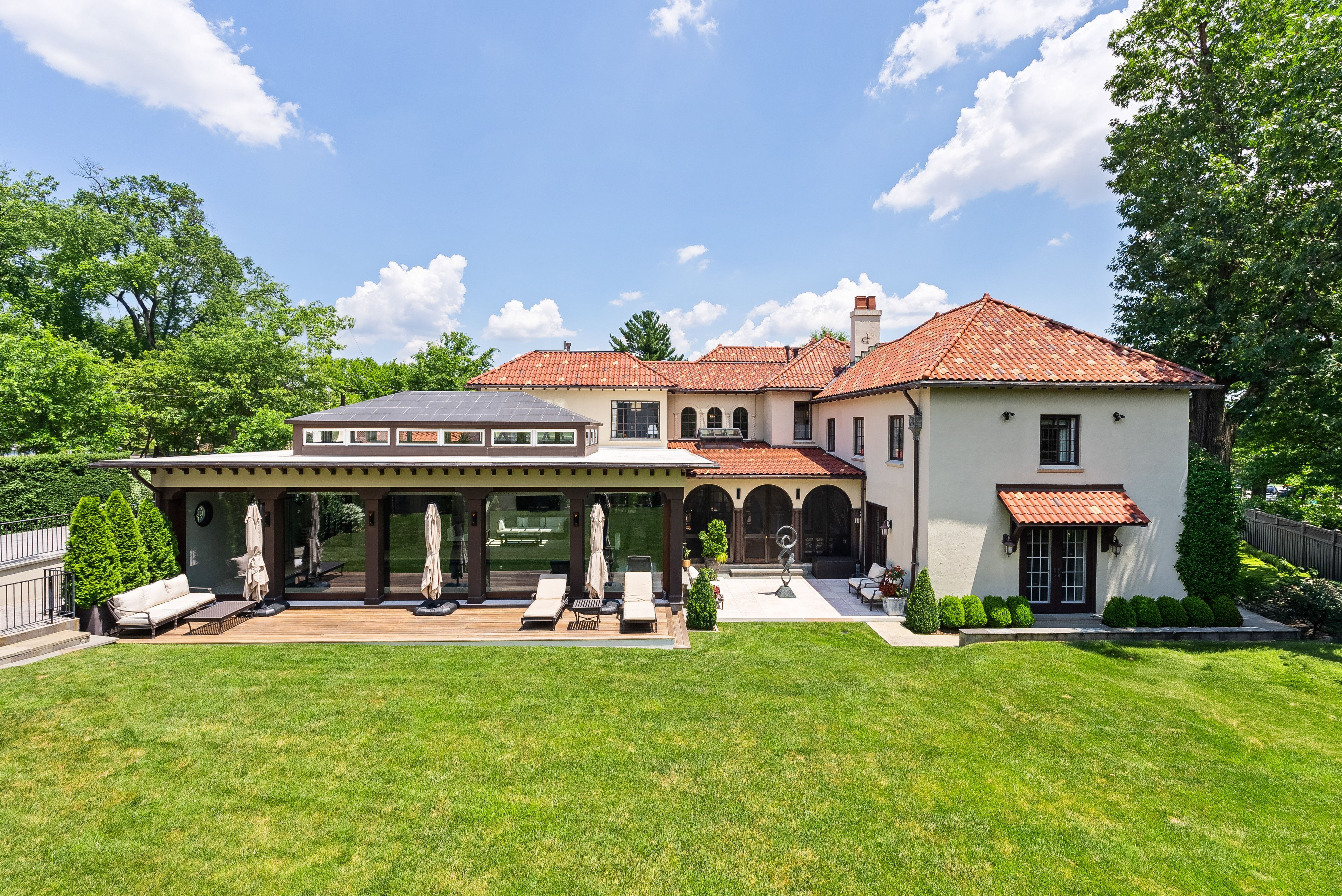 Two-story Mediterranean-style home with red-tiled roof and beige stucco, glass-walled patio, lounge chairs and umbrellas on a wooden deck, green lawn, tall trees, and a bright blue sky.