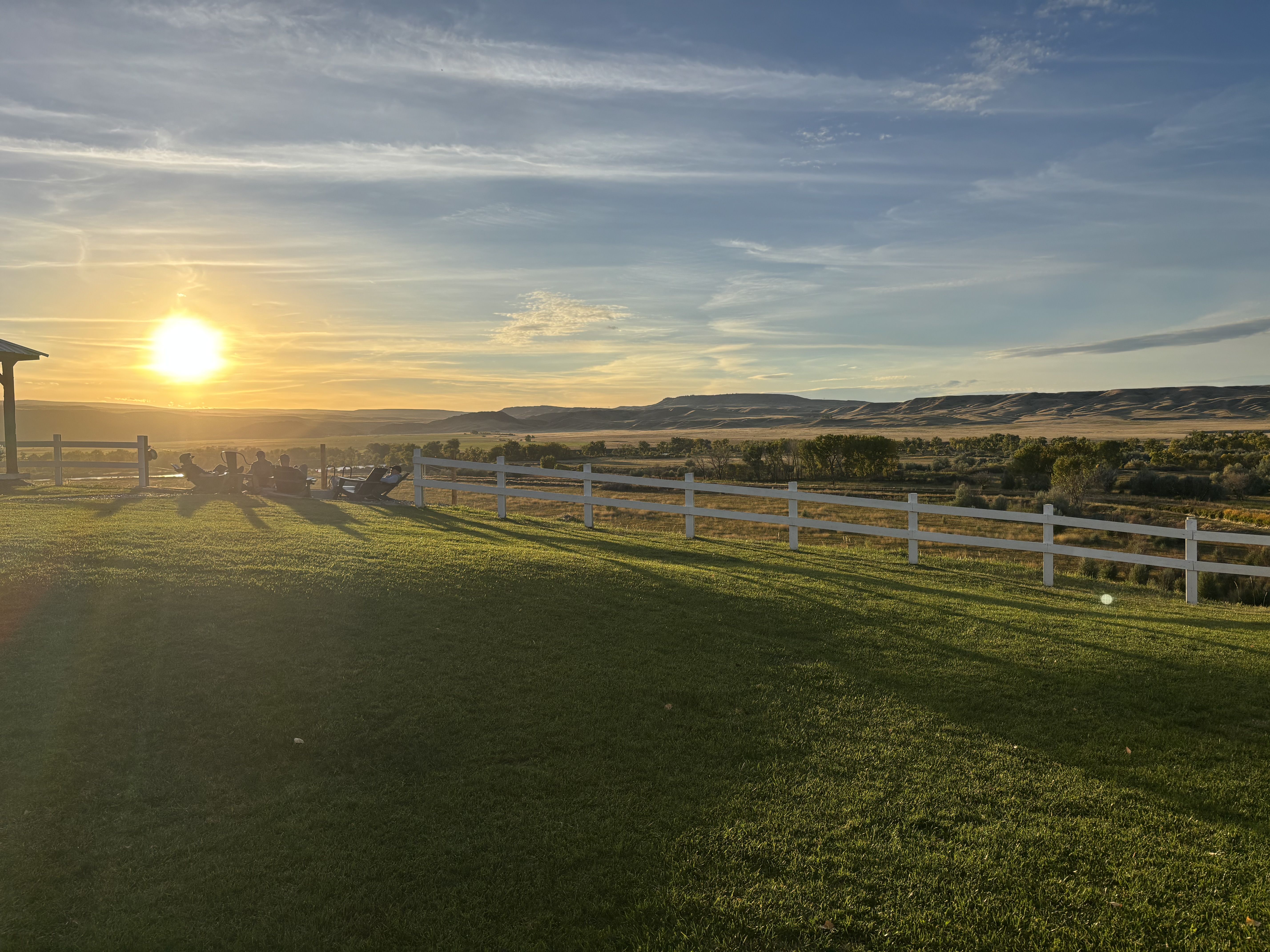 Golden sunset over a rural field with a white fence, distant hills, and a few people sitting by the fence on the left. Bright sun near the horizon casts long shadows.