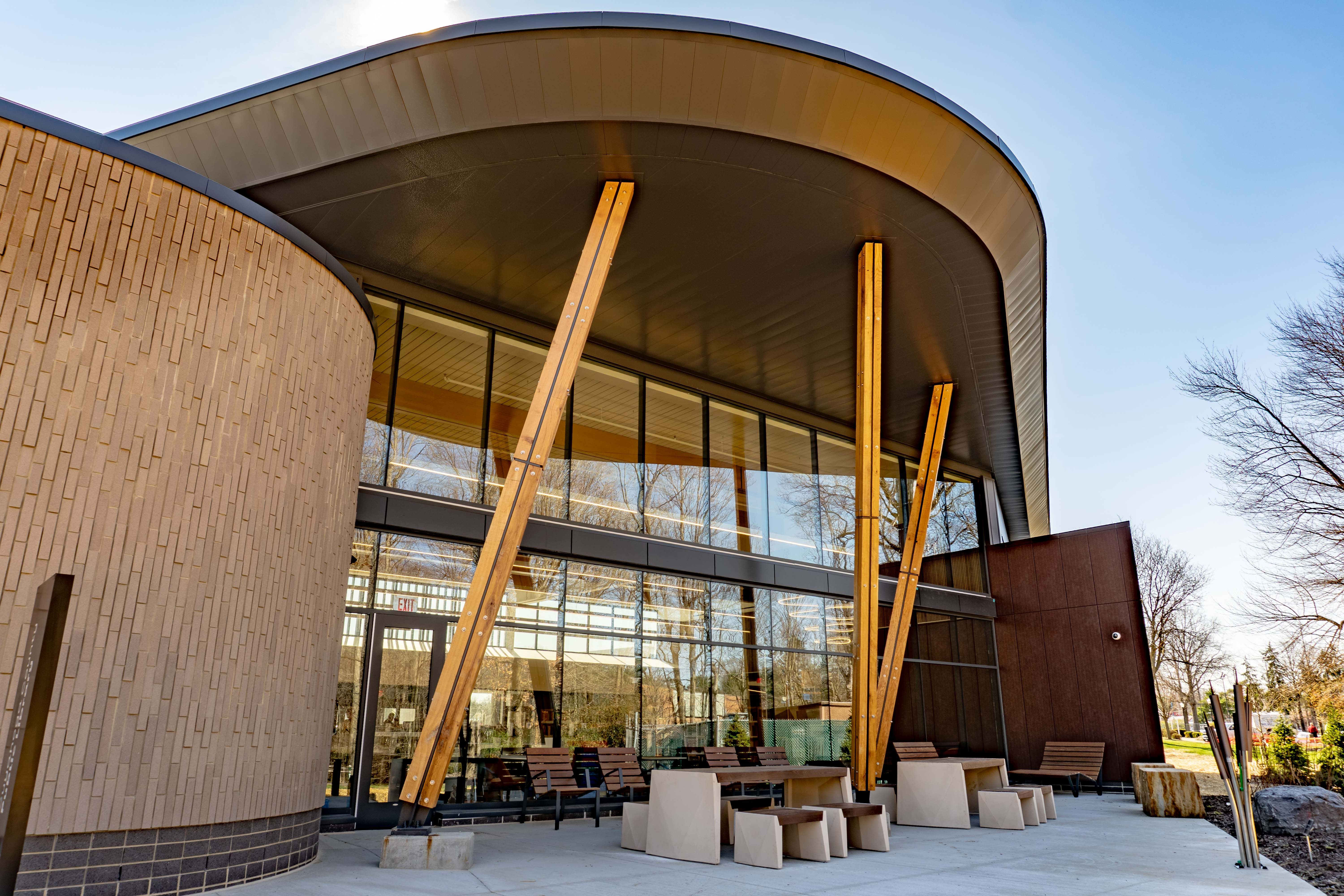 Modern building with a curved metal roof and large glass walls. A rounded brick wall on the left, two tall wooden braces support the overhang; concrete benches sit outside under a blue sky.