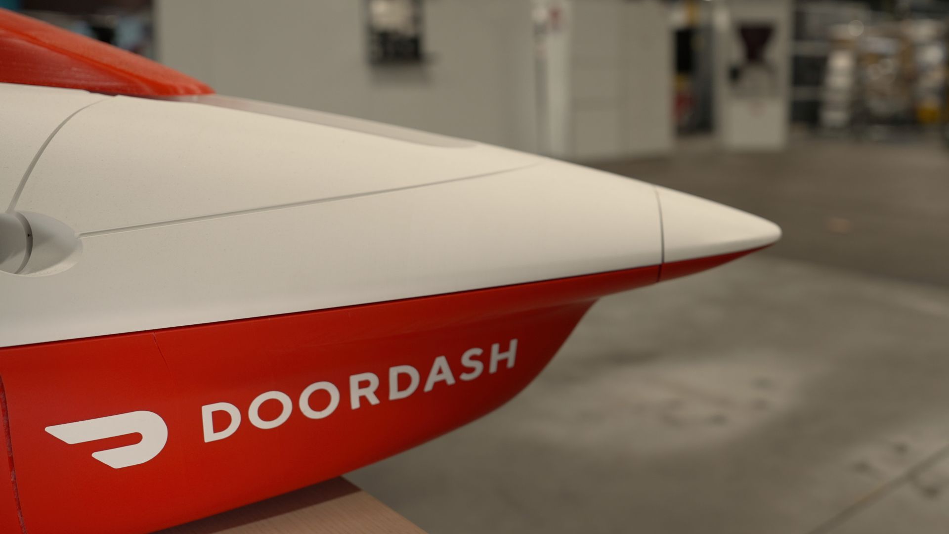 Close-up of a white and red delivery drone with DOORDASH on its red fuselage, indoors in a hangar with blurred equipment in the background.