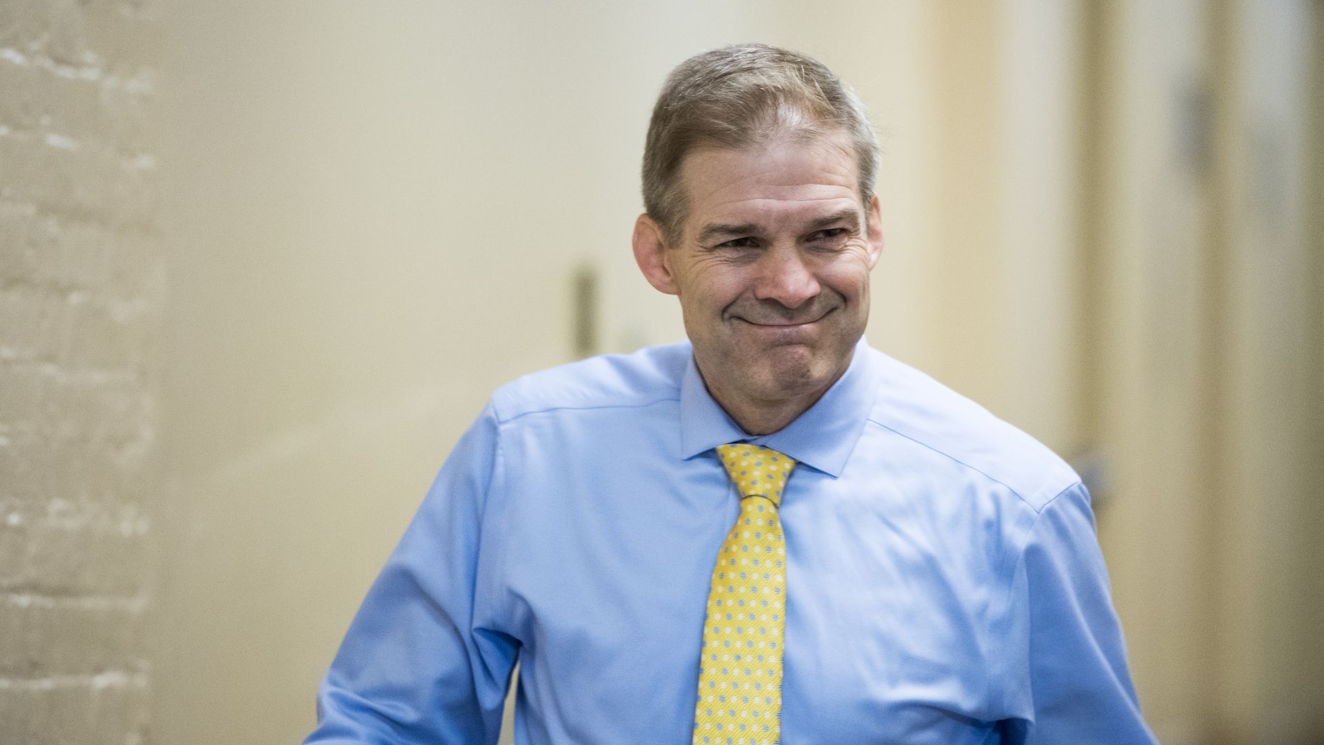 Rep. Jim Jordan in a blue shirt and yellow tie smiles in a strained way while walking.