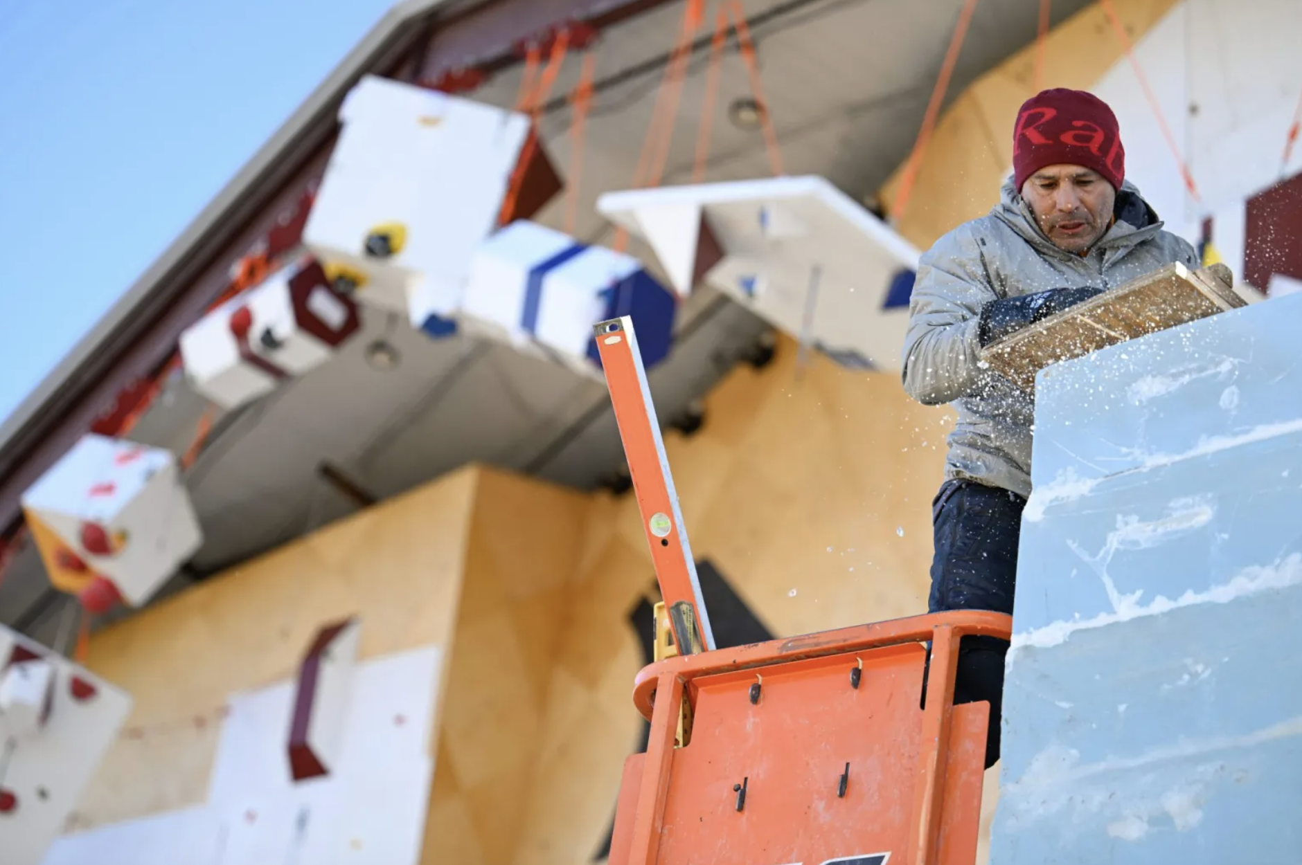 Man wearing a gray jacket and red beanie carves a large blue ice block with a tool, standing on an orange platform, with geometric colored blocks hanging in background.