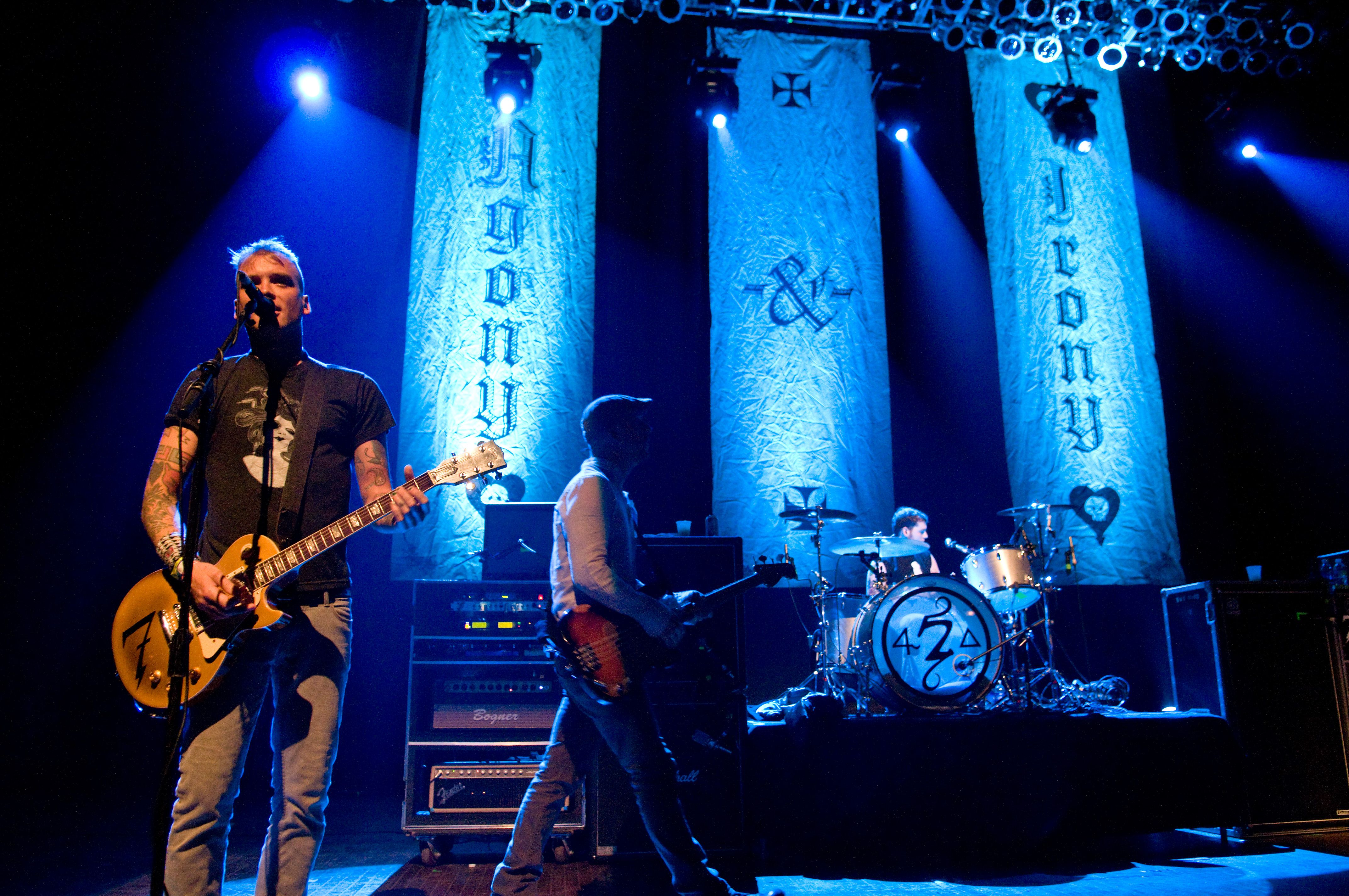 Blue-lit rock concert: a guitarist in a black T-shirt plays a sunburst electric guitar at the mic, with a drummer, amplifiers, and tall textured banners behind on stage.