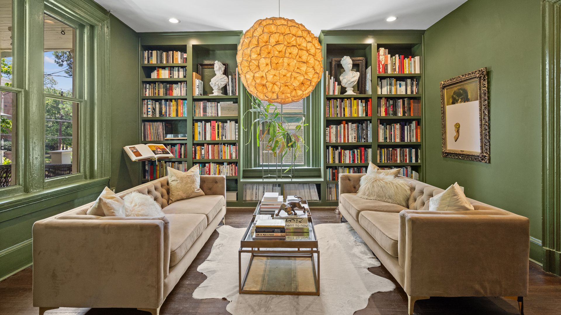 Two beige couches face each other in a green library with a bold orange chandelier hanging from the ceiling and a furskin rug on the hardwood floor.