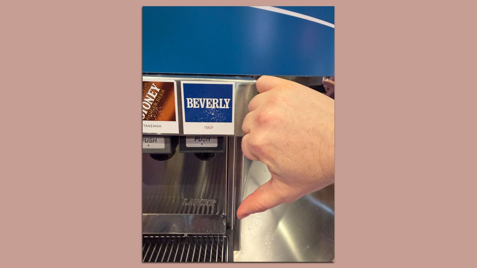 Close-up of a hand giving a thumbs down in front of a soda fountain; labels show brown "Tanzania" and a blue "Beverly" option on a shiny stainless machine with a blue top.