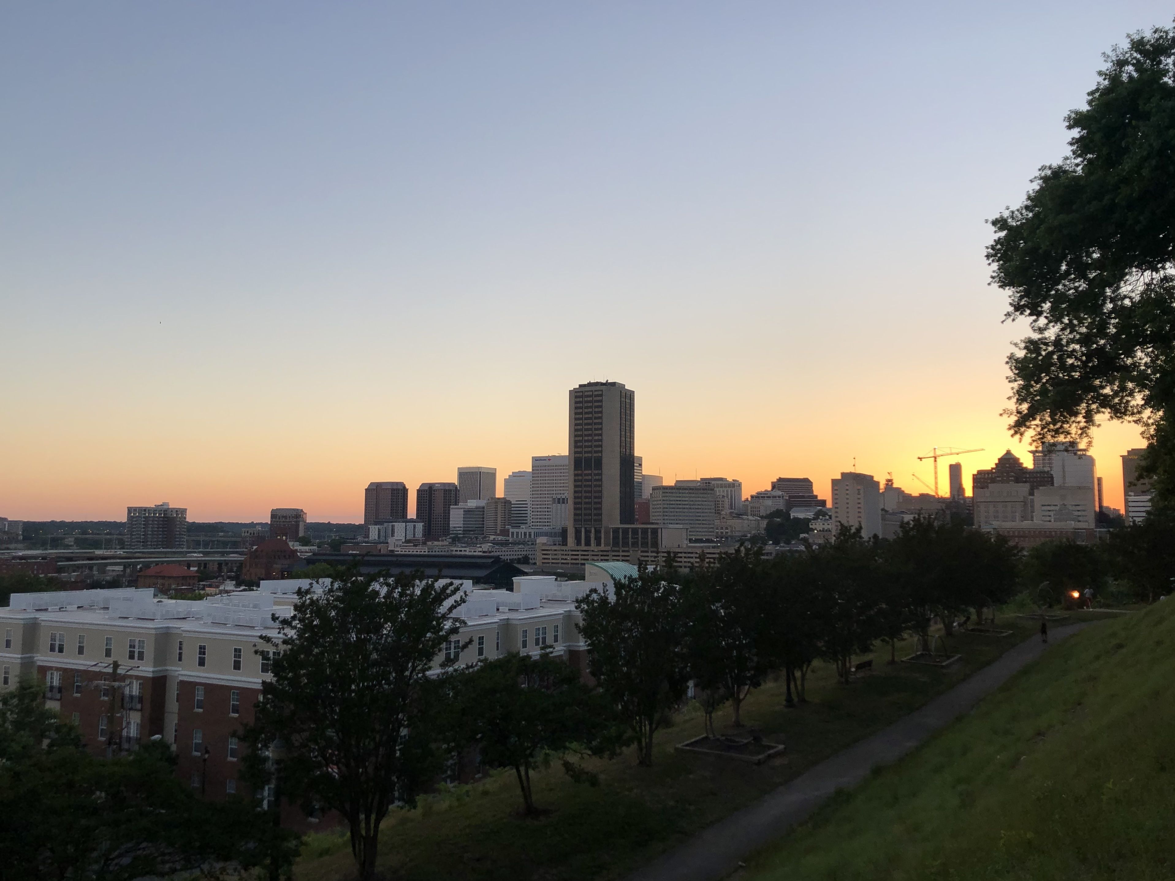 City skyline at sunset with orange and purple sky, tall buildings, trees, and a walking path on a grassy hill in the foreground.