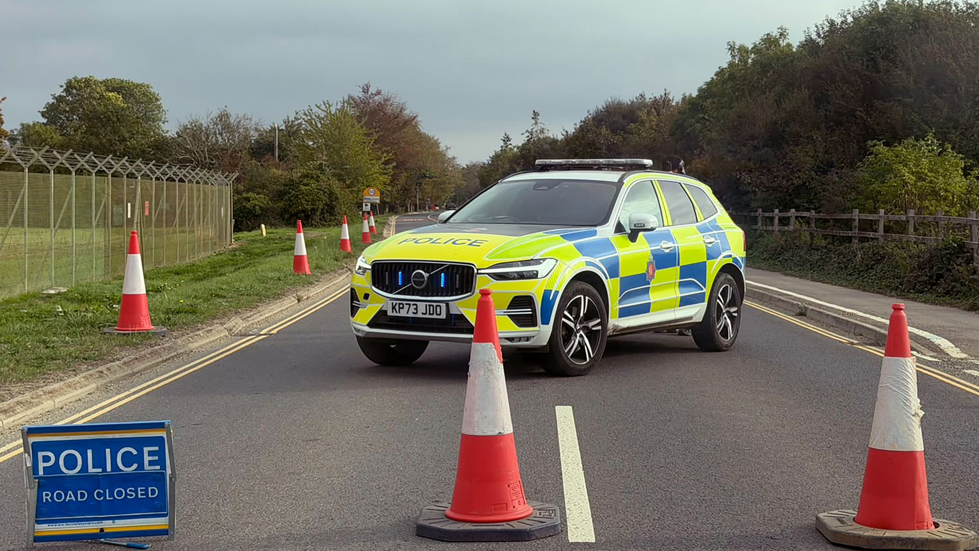 Police SUV with bright livery blocks a road; orange cones line the lane; foreground shows a "POLICE ROAD CLOSED" sign, with fencing and trees along the road under a cloudy sky.