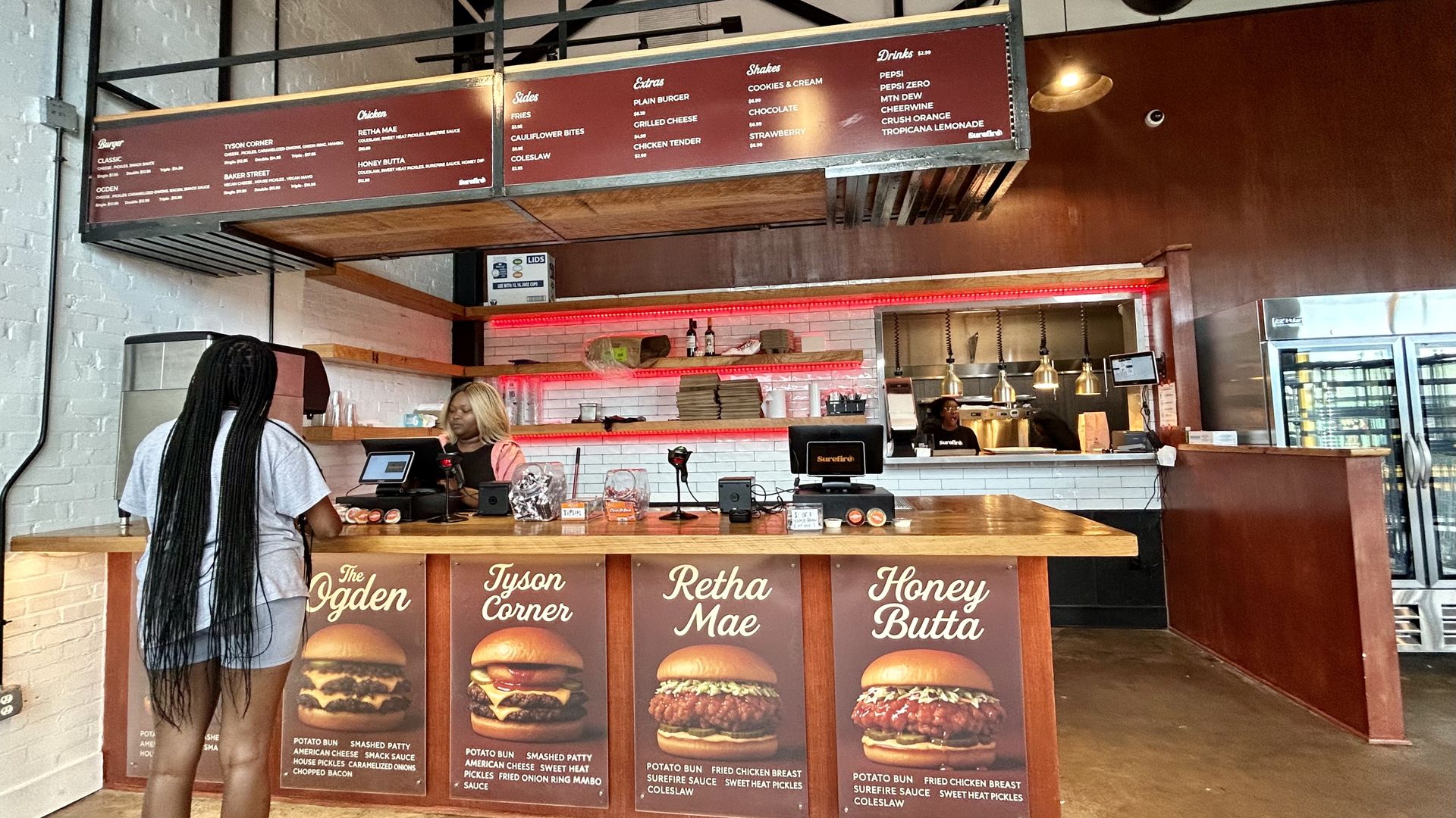 Interior of a burger restaurant with red menu boards listing burgers, sides, shakes, and drinks. A customer with long braids stands at the counter where a worker takes an order.