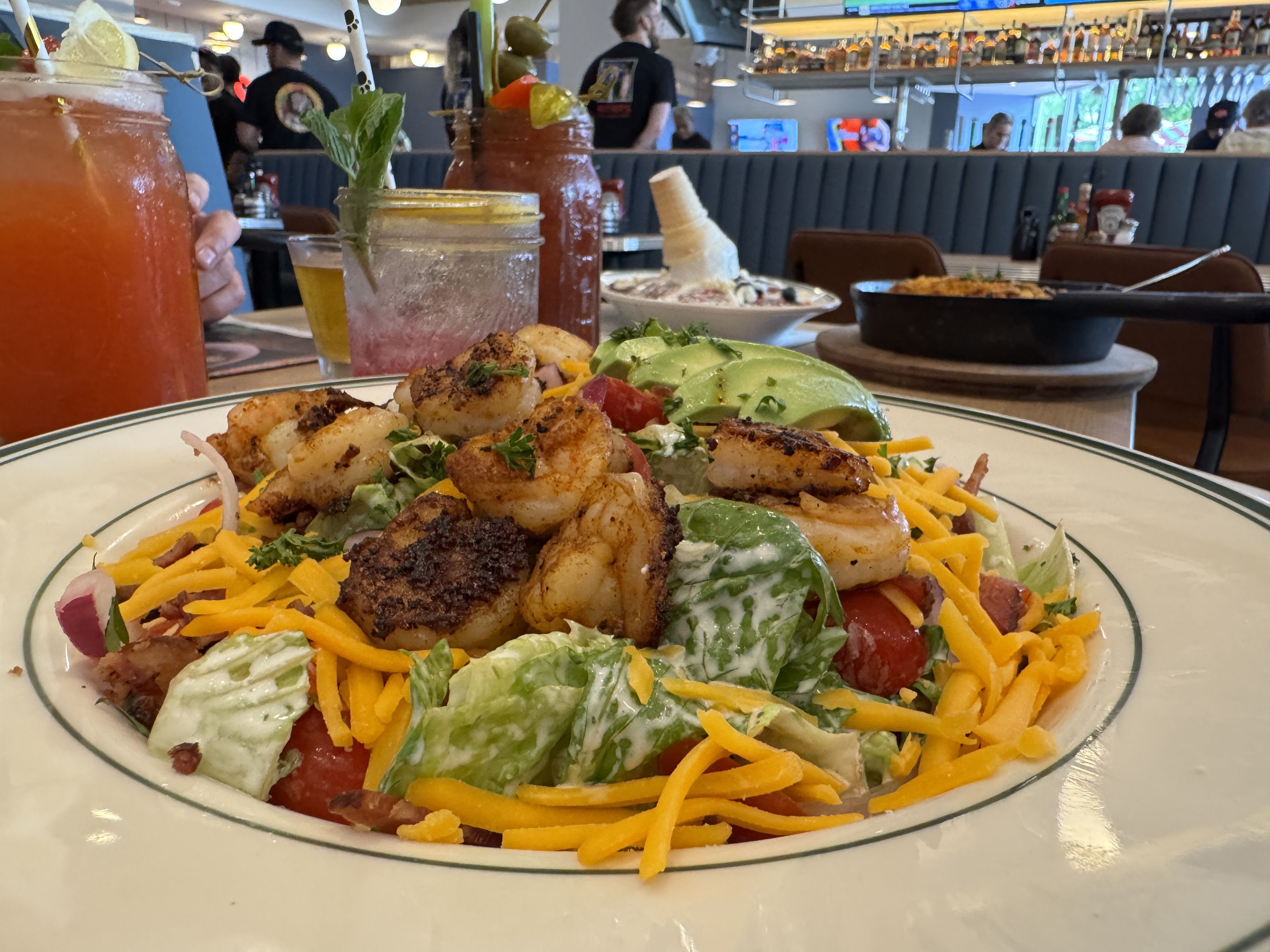 Grilled shrimp salad with lettuce, shredded cheddar cheese, avocado slices, and cherry tomatoes on a white plate in a busy restaurant with drinks in the background.