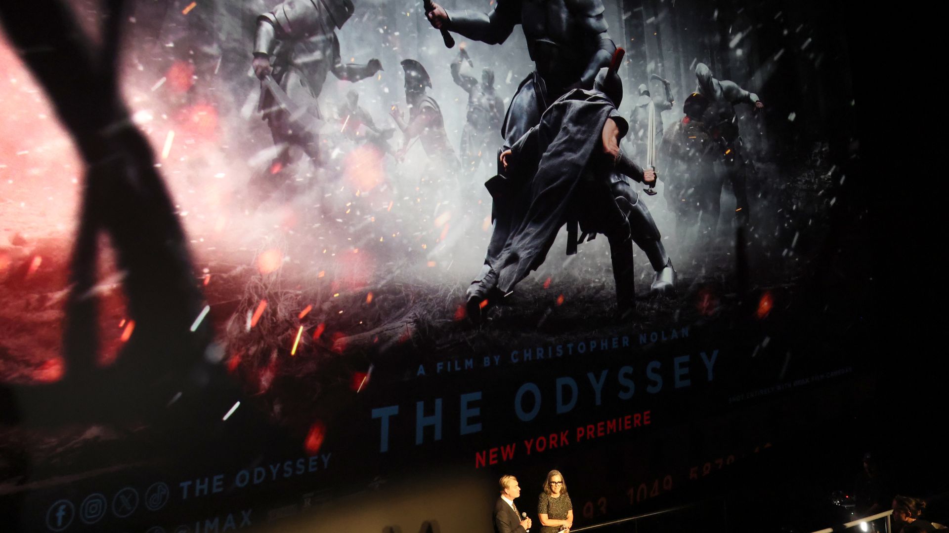 Movie screen showing soldiers fighting in "The Odyssey" and two people stand on stage during the New York premiere