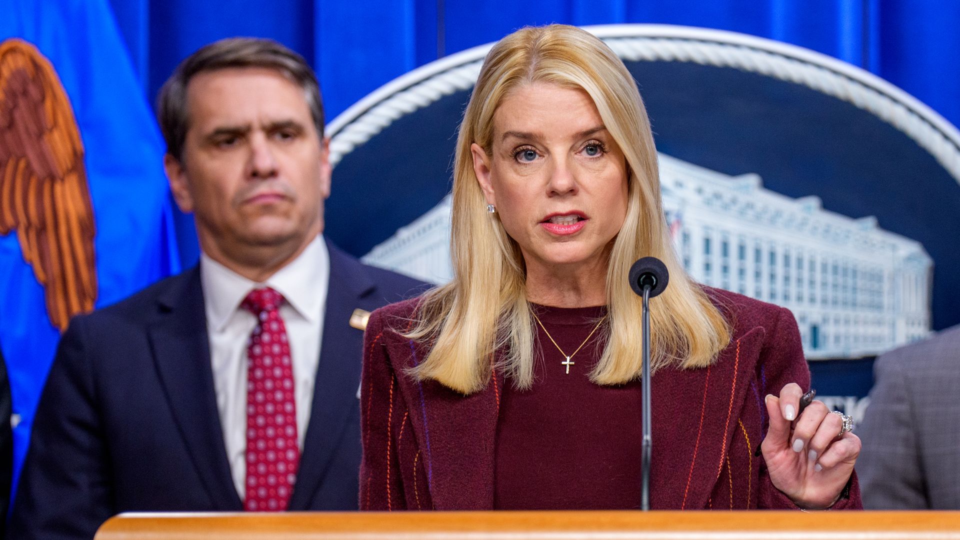Three officials stand at a Department of Justice podium with a seal; a blonde woman in a maroon jacket speaks into a microphone, flanked by two men in suits and red patterned ties.