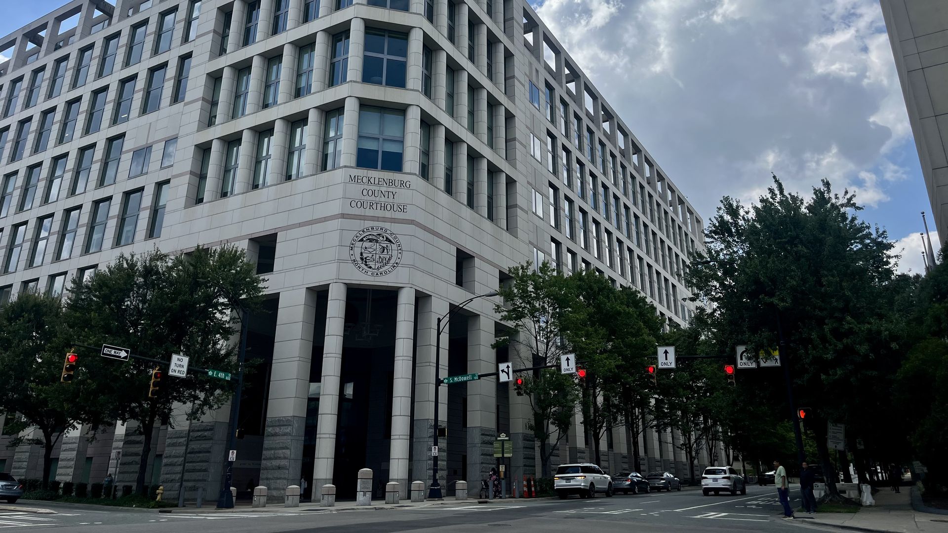 The Mecklenburg County Courthouse, a modern multi-story building with white and gray exterior, seen at an intersection with traffic lights and trees under a partly cloudy blue sky.
