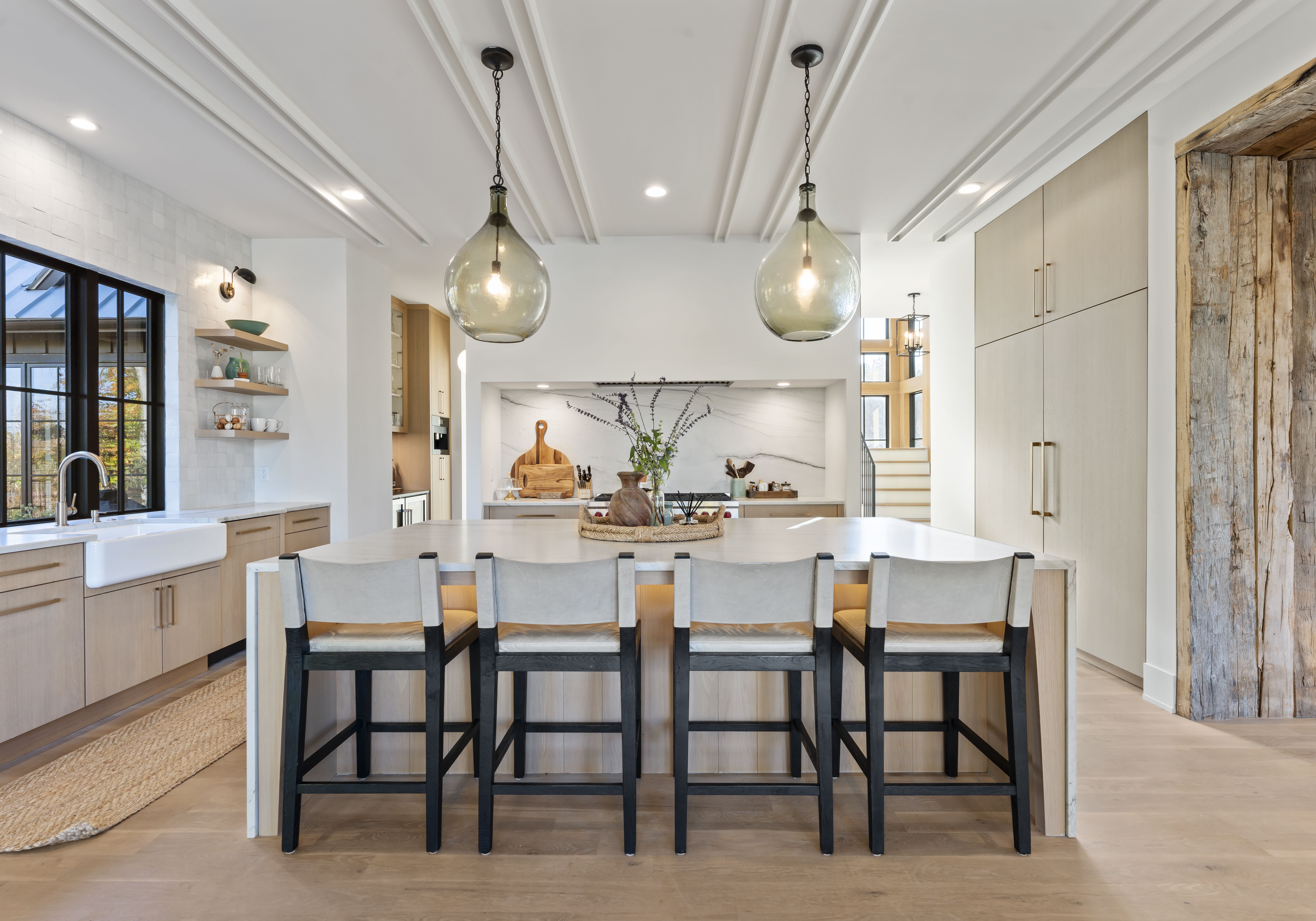Bright modern kitchen with a large white island and five light stools with dark legs. Two glass pendant lights hang from a paneled ceiling, with light wood cabinets and a rustic timber post.