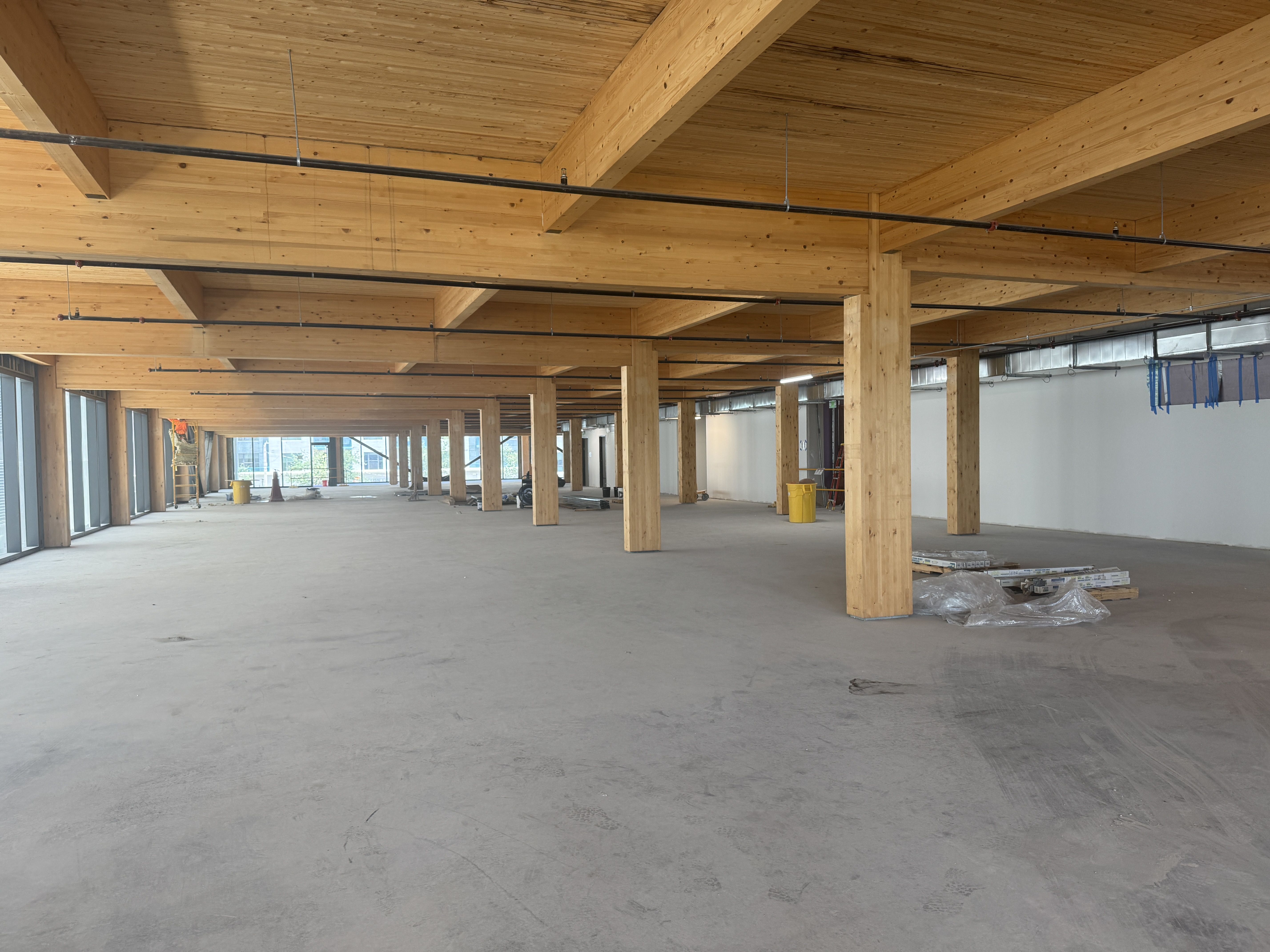 Interior of a large unfinished building with exposed wooden beams and columns, black pipes along the ceiling, a concrete floor, and scattered construction materials and yellow bins.