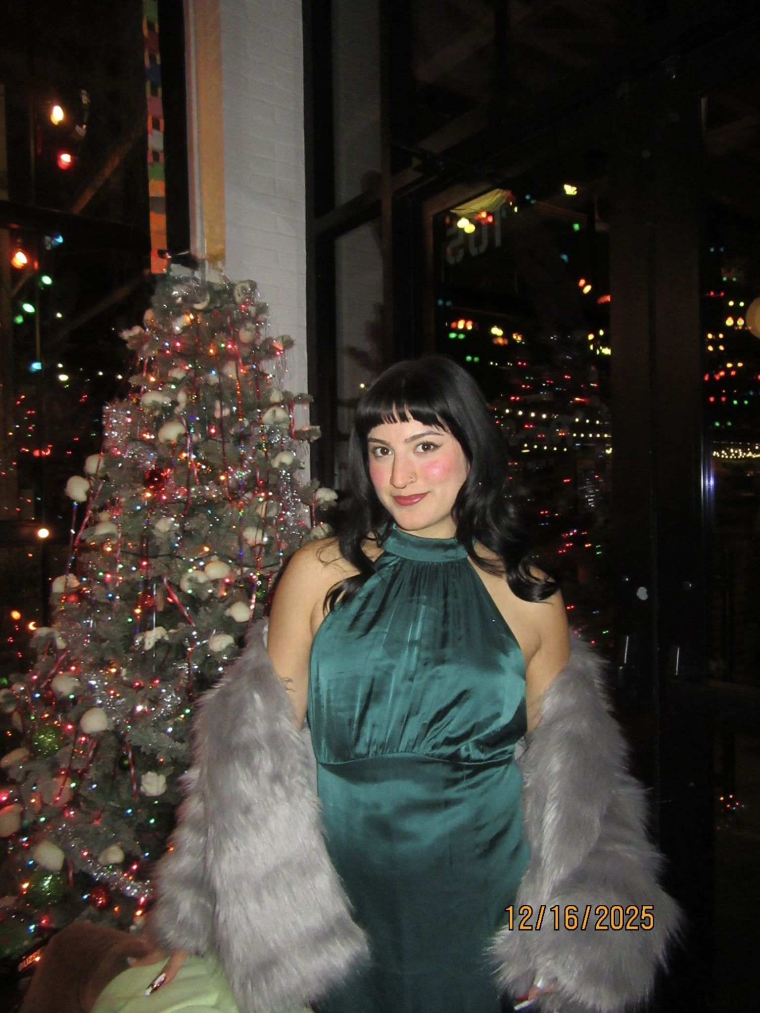 Woman with black hair in a teal satin halter dress and gray fur coat stands beside a lit Christmas tree; colorful city lights glow in the background, with a date stamp reading 12/16/2025.