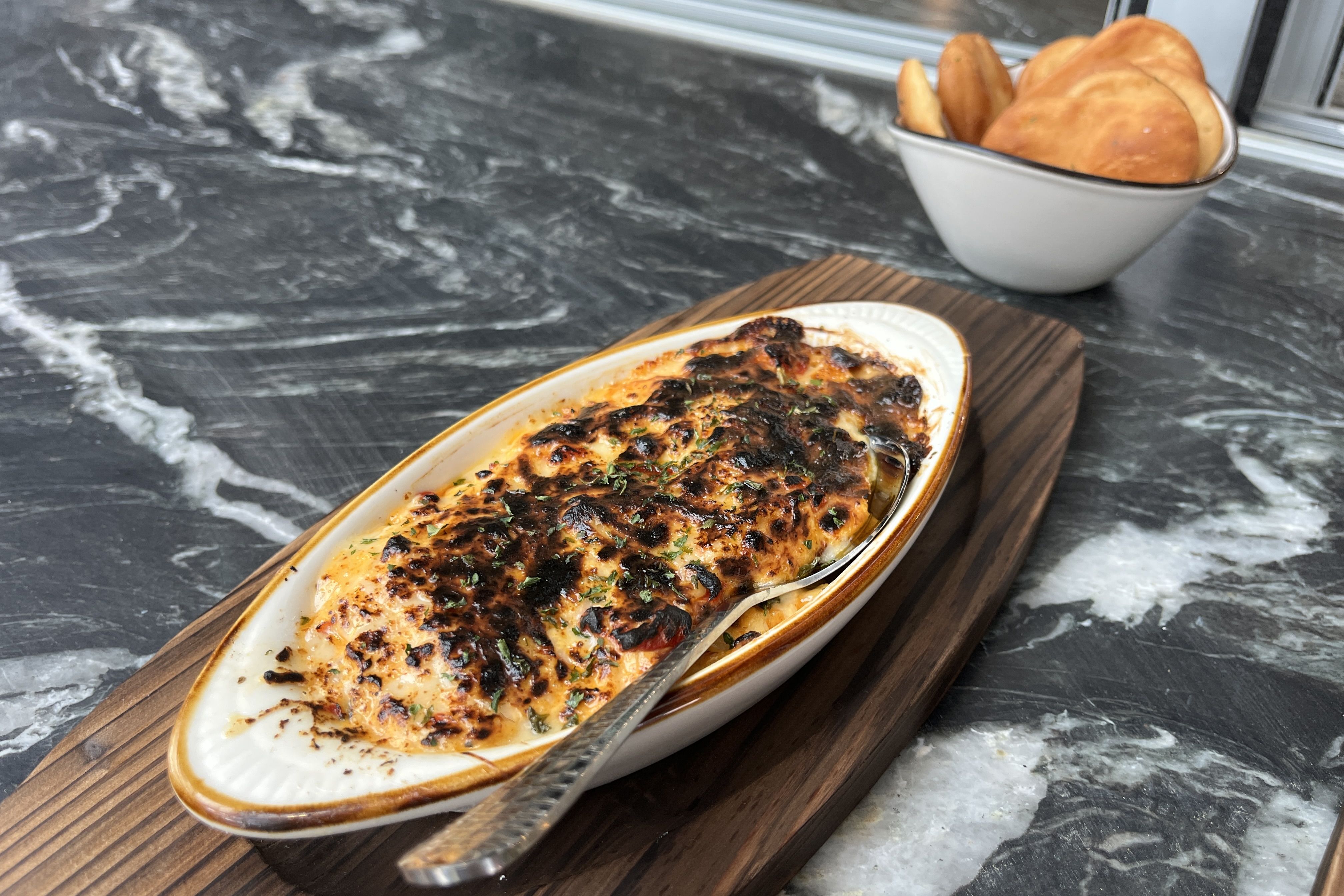 A baked cheesy casserole with a browned top in a white oval dish sits on a wooden cutting board; a white bowl of golden bread rolls rests on a dark marble countertop.