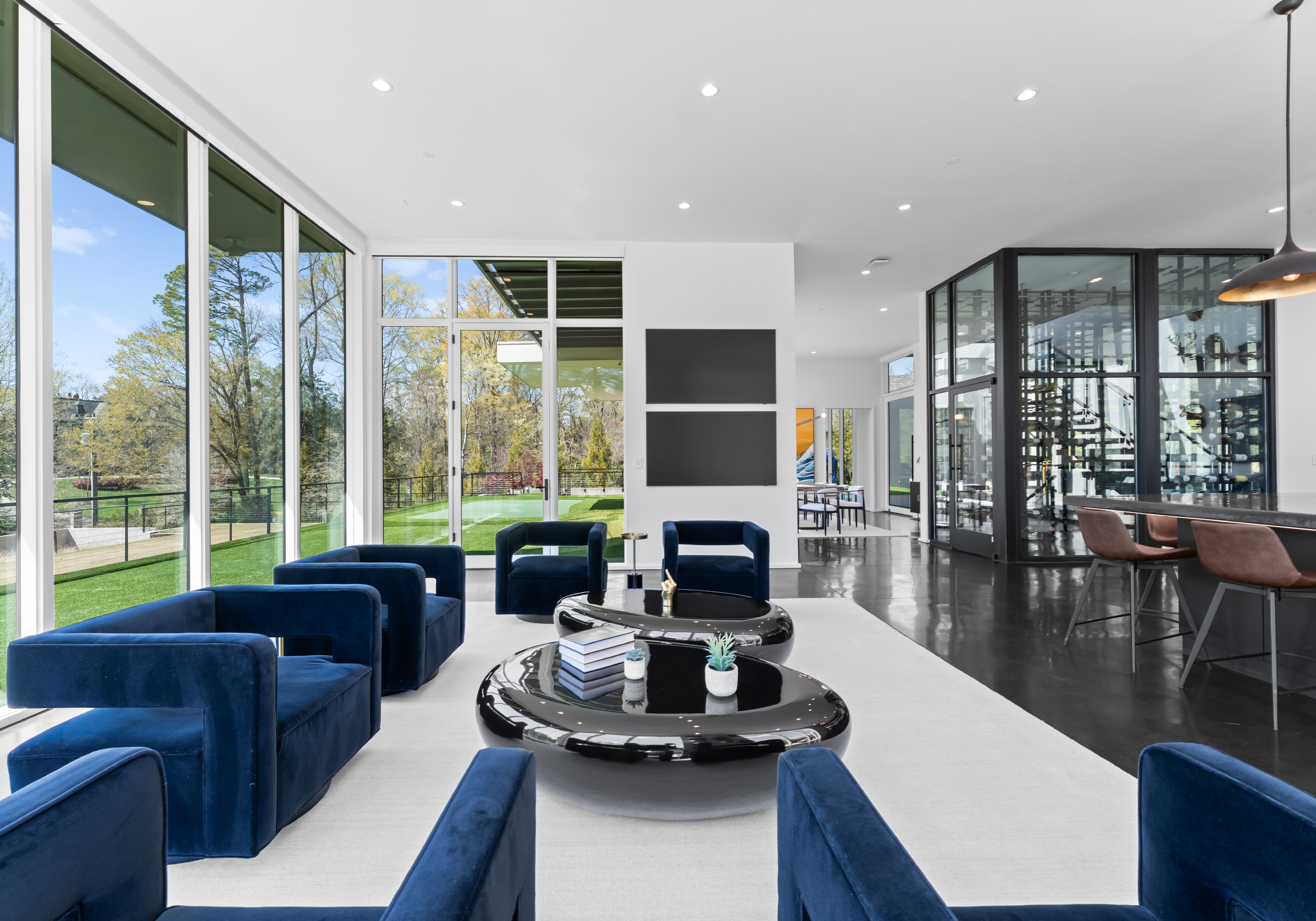 Modern living room with large glass windows, blue velvet chairs, black glossy ottomans, white rug, and a view of green lawn and trees outside under blue sky.