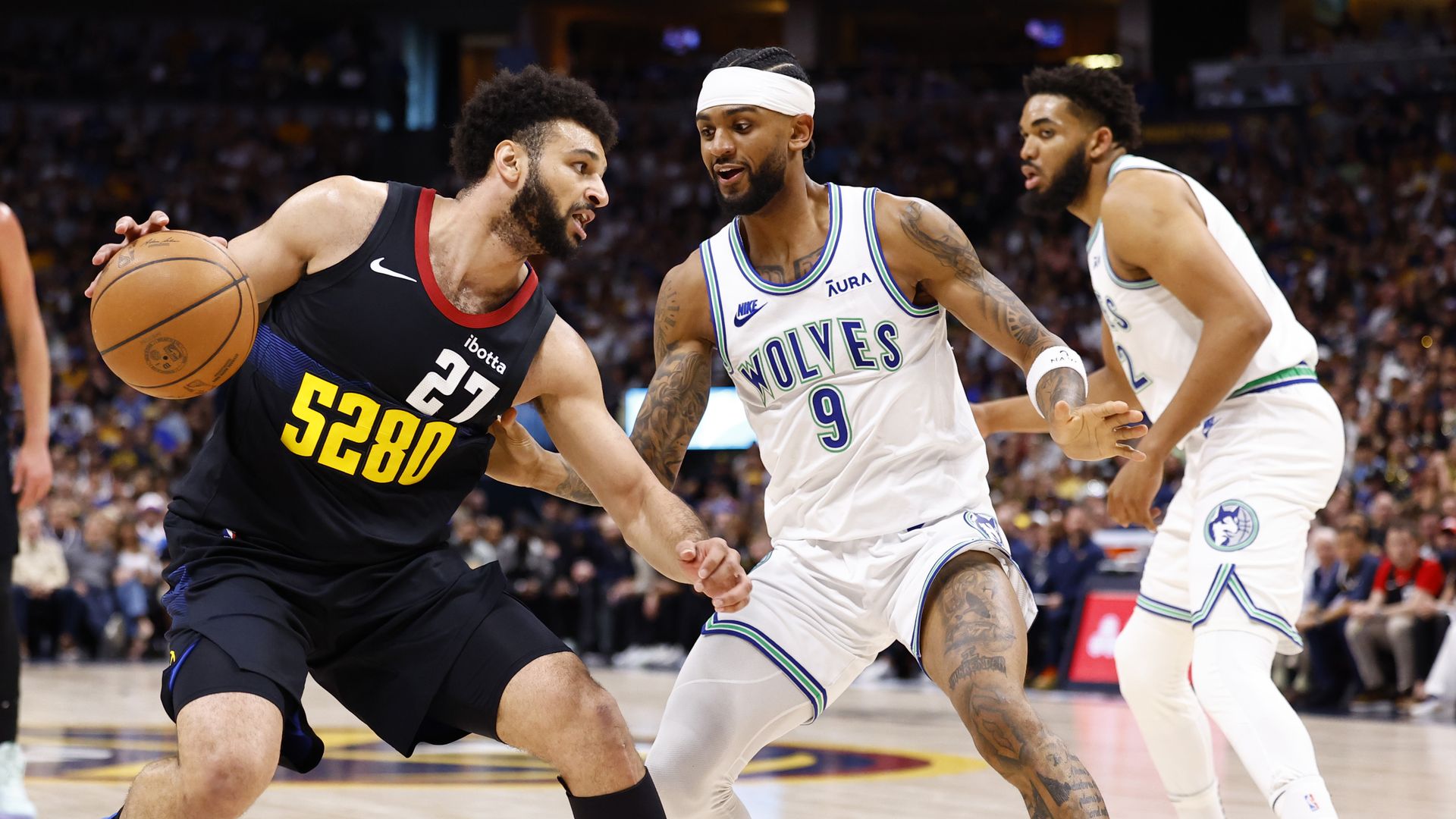A man with a black jersey with the number 5280 and 27 dribbles an orange basketball while two players in white jerseys, including one with the words WOLVES on it, during a game.