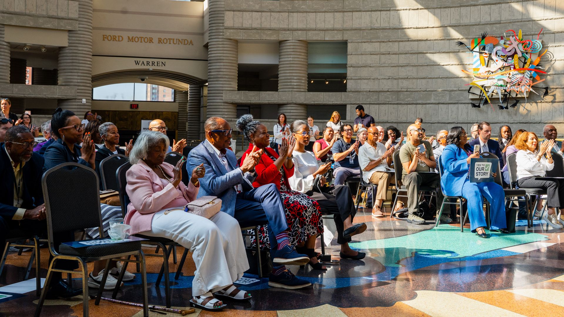 Audience seated and clapping in a sunlit atrium with stone walls and large columns.
