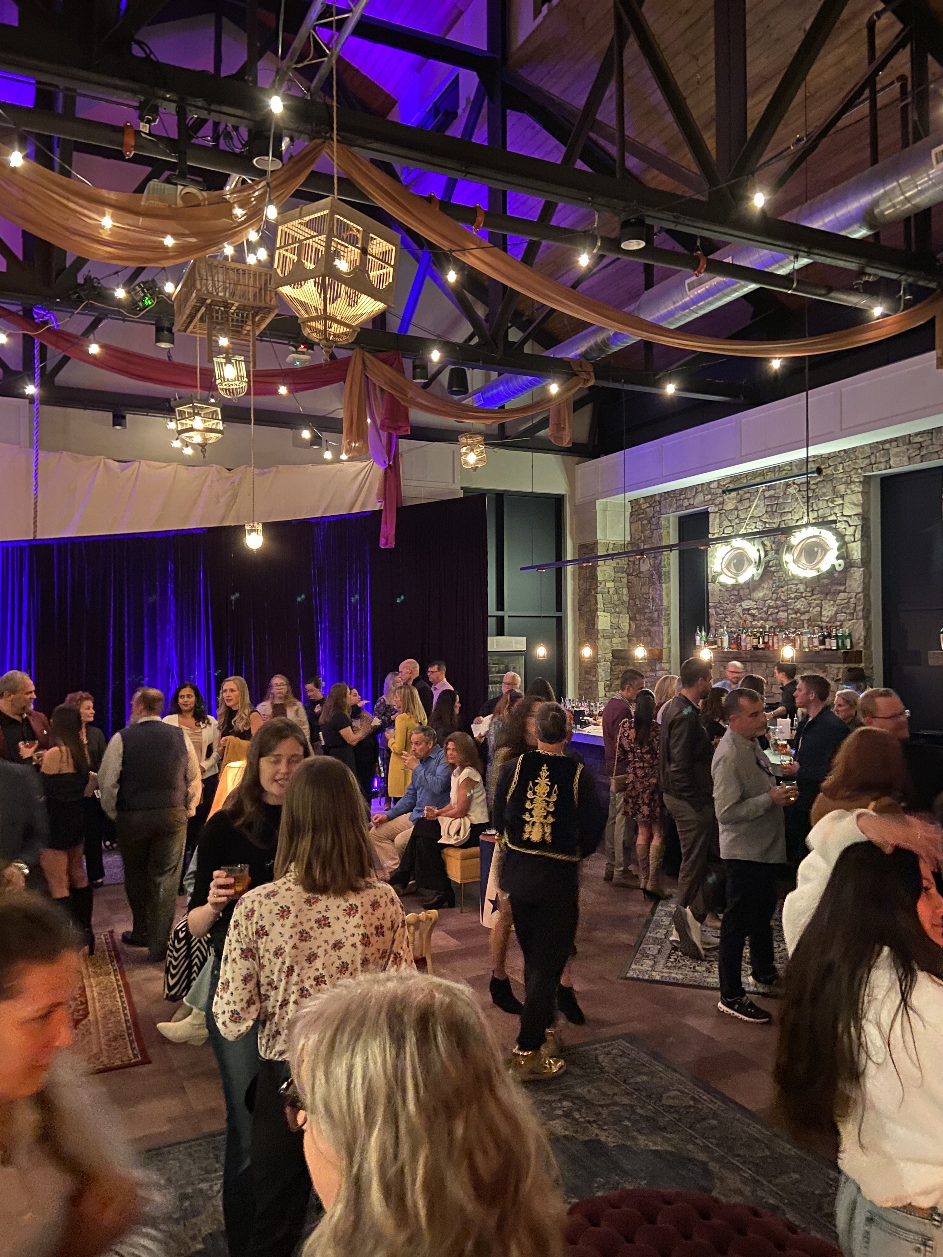 Indoor party with diverse crowd socializing under warm string lights and draped orange fabric, with glowing eye art on exposed brick wall and purple stage curtains in background.