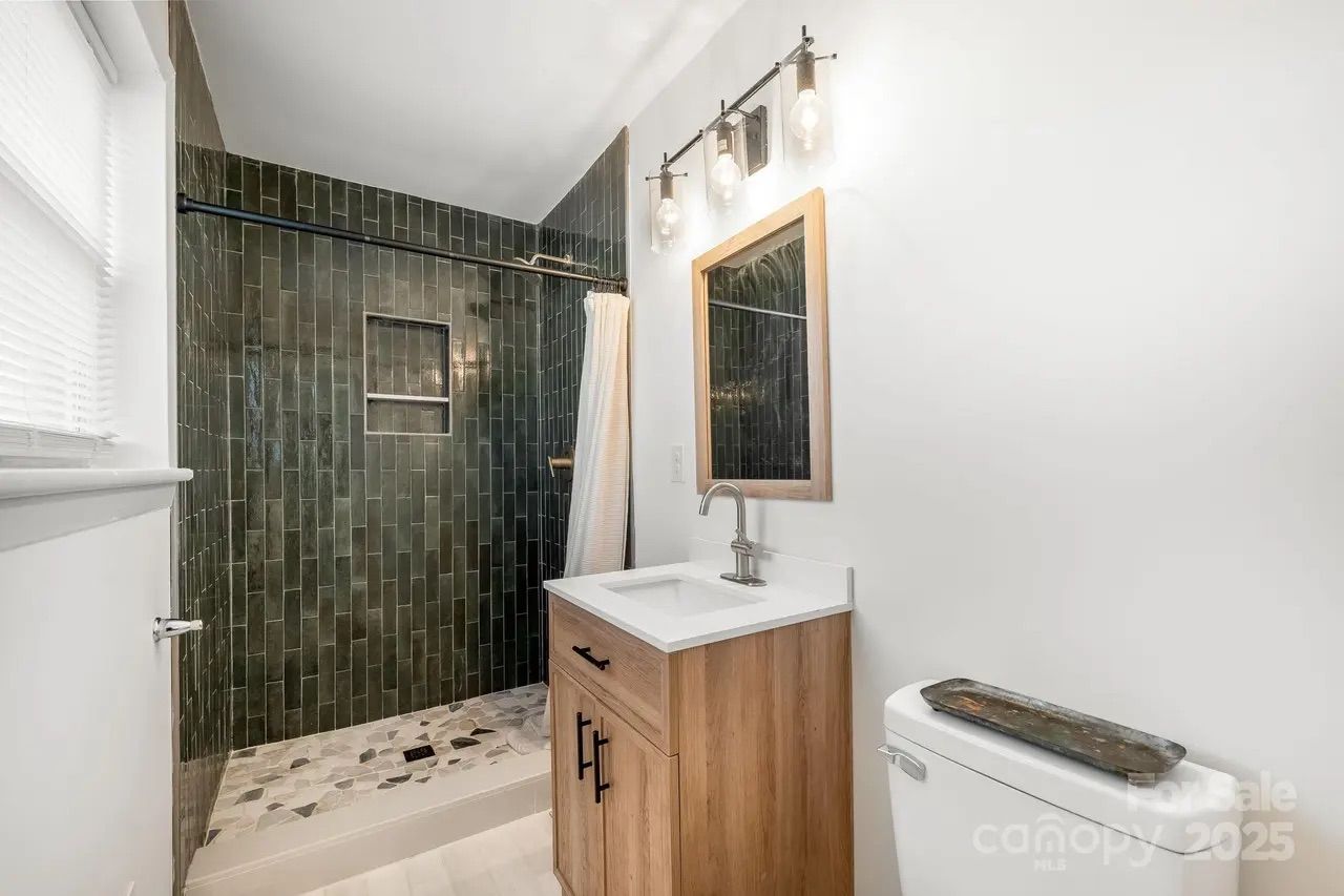 Modern bathroom with dark green vertical tile shower, white countertop vanity with wooden cabinet, rectangular mirror, three-bulb light fixture, and white toilet with decorative tray.
