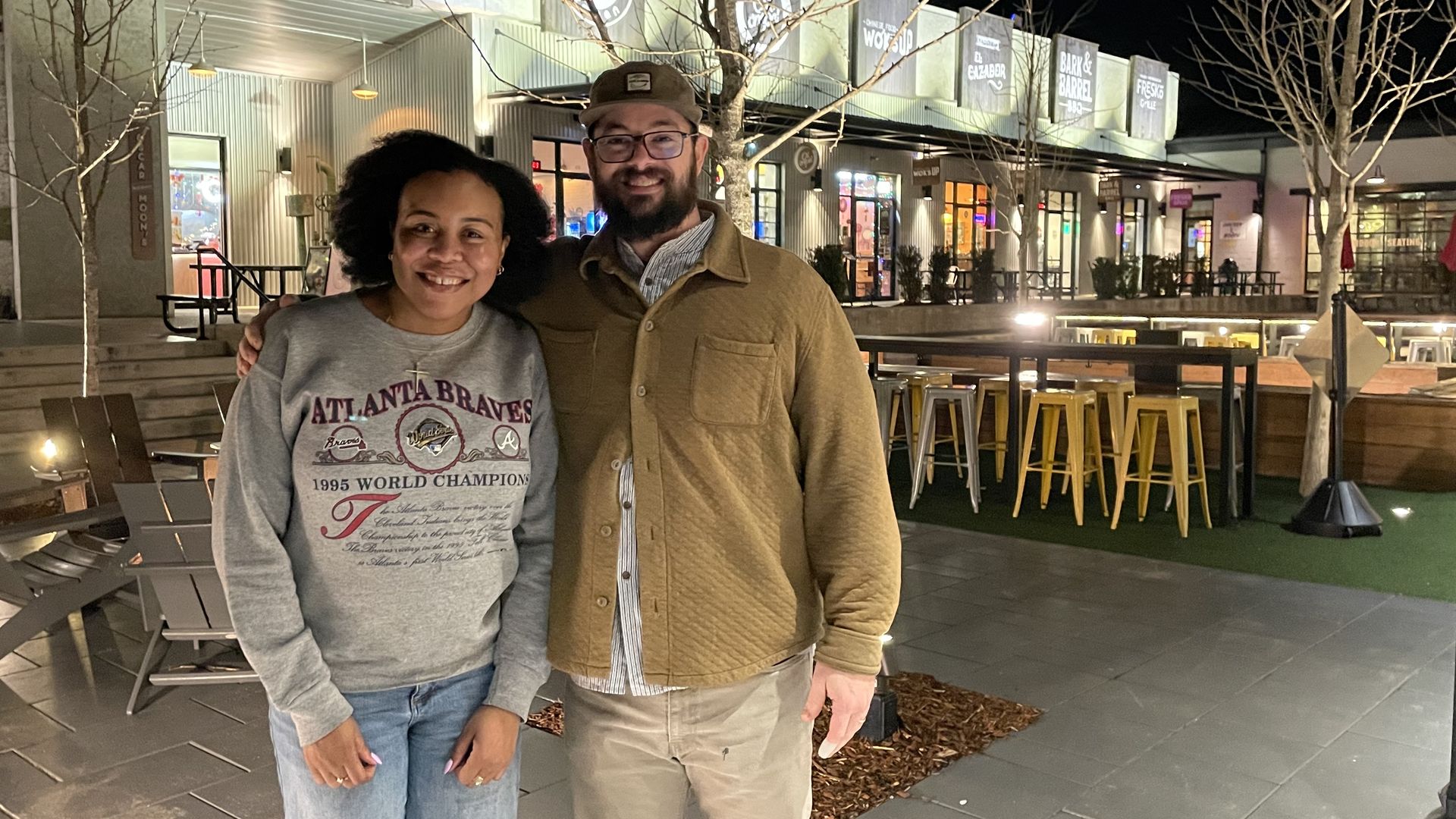 Two people smiling outdoors at night in front of a shopping area with illuminated signs, bare trees, yellow stools, and patio seating. One wears an Atlanta Braves sweatshirt; the other wears a brown jacket.