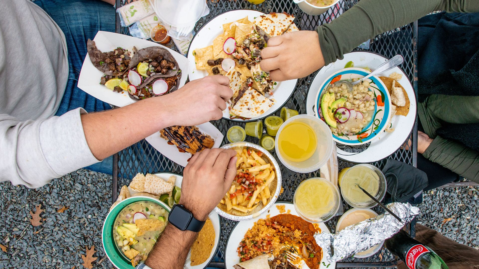 An aerial photograph of hands reaching for colorful plates of Mexican food crowding a small table