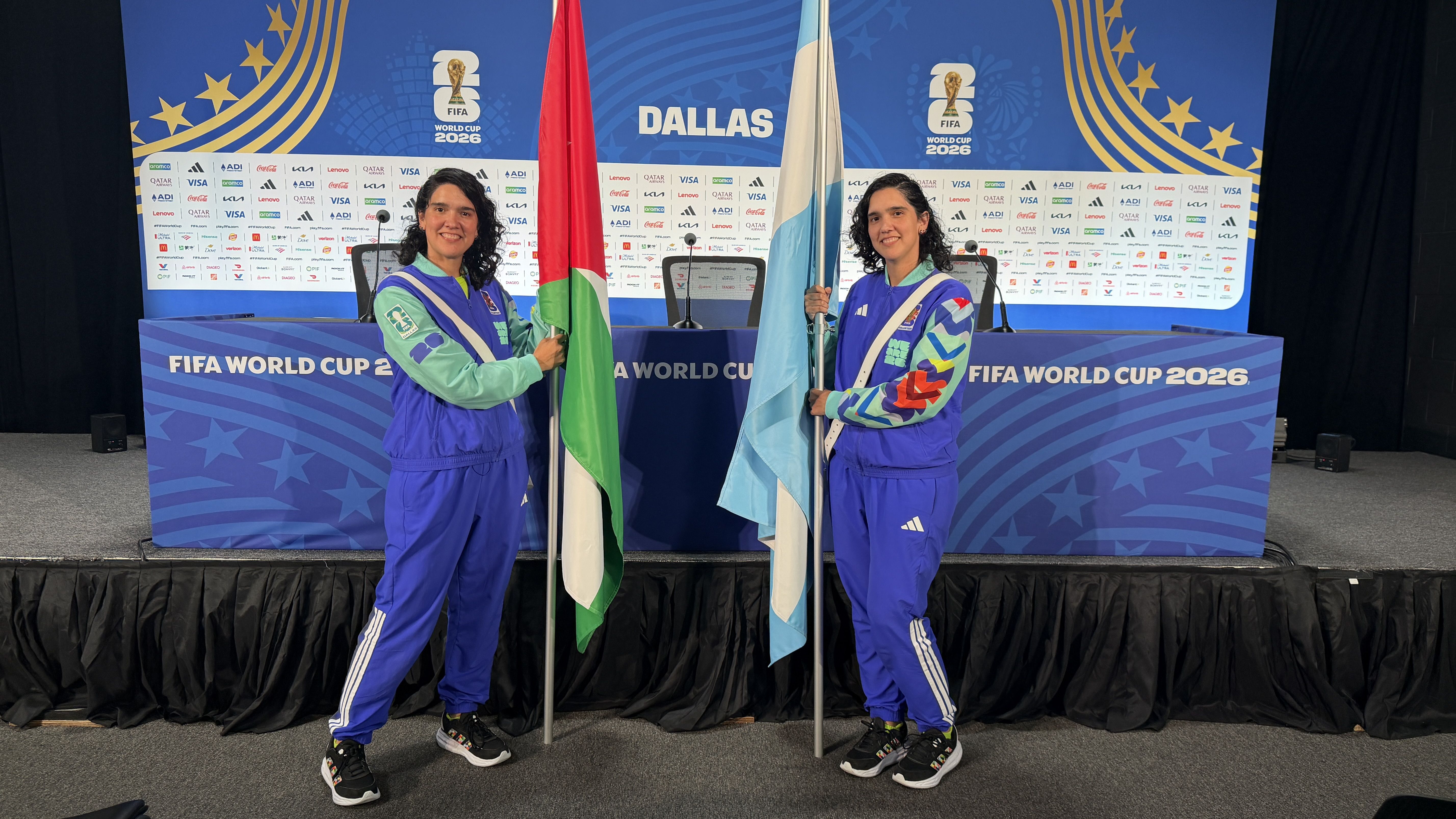 Two women in blue athletic outfits stand on a press stage, each holding a flagpole with a flag. Behind them is a blue Dallas backdrop with FIFA World Cup 2026 and sponsor logos.