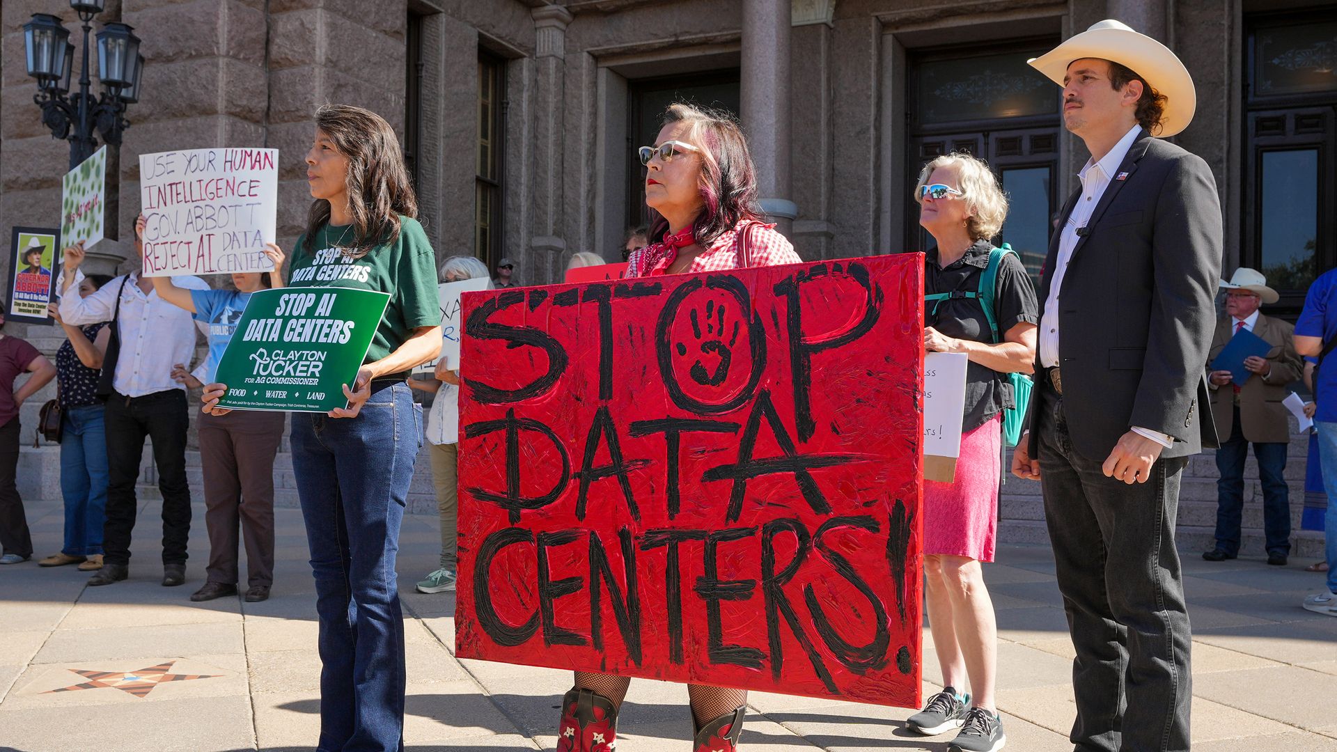 A rally at the Texas Capitol protesting data center expansion before a Senate committee hearing in July. A woman holds a large red sign that reads, in black text, "STOP DATA CENTERS!" Next to her stands a woman holding a smaller green sign that reads "Stop AI data centers."