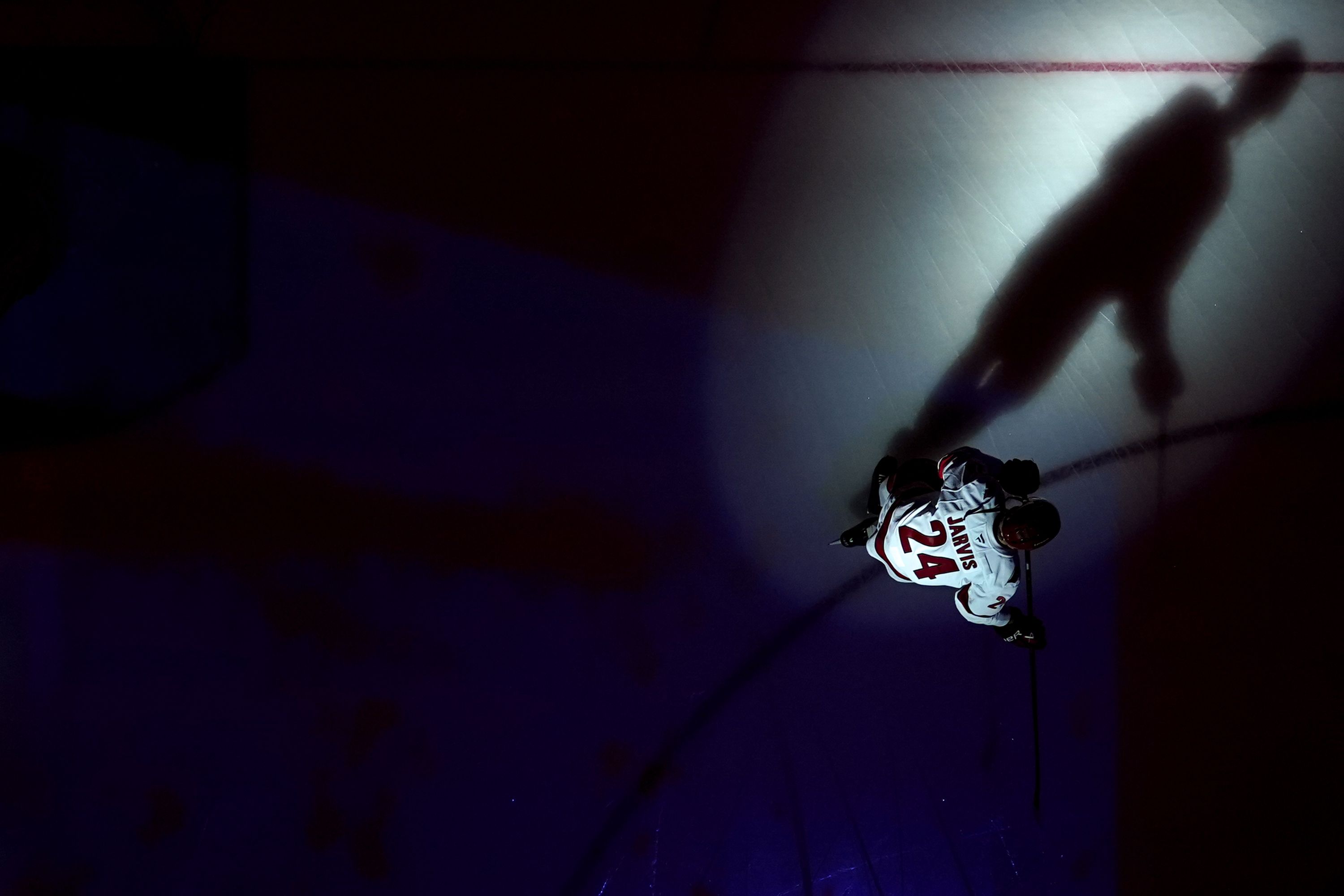 A Carolina Hurricanes player wearing a white jersey kneels alone under a spotlight on the dark ice, his shadow stretching across the rink before a game.