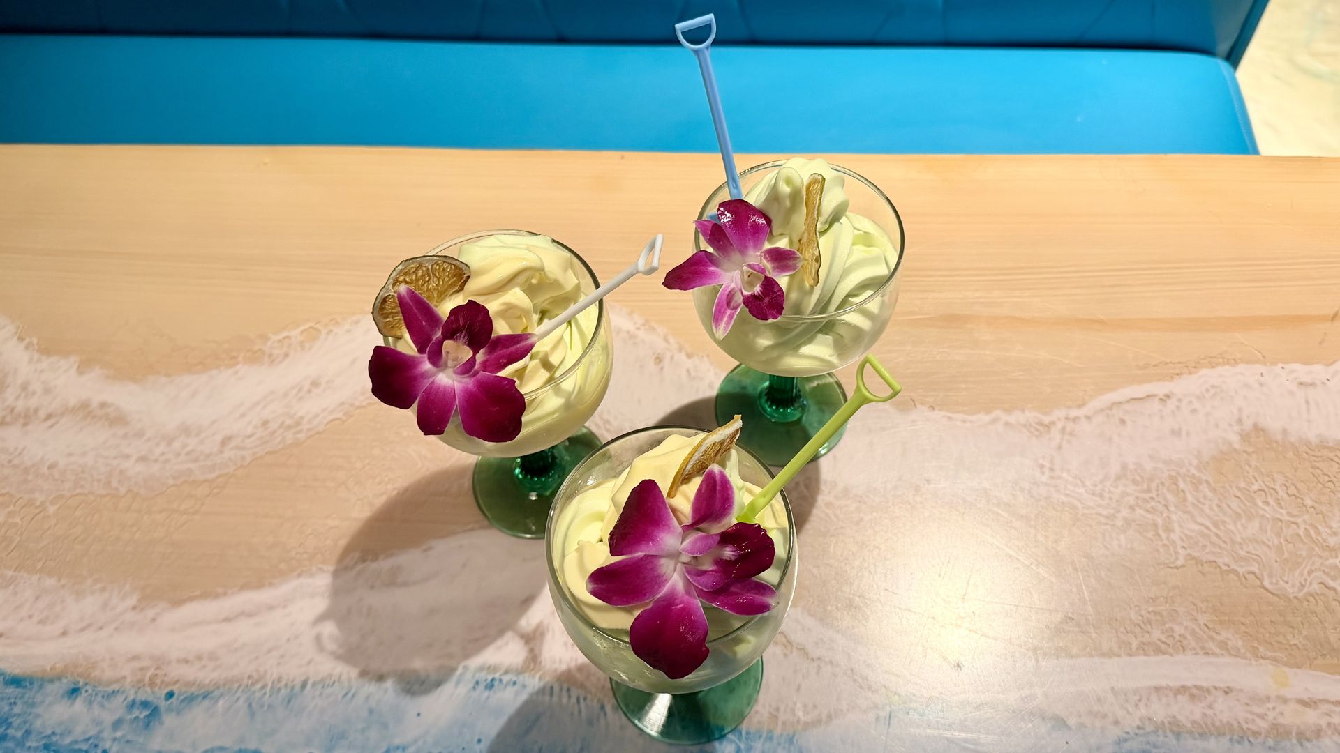 Three tropical cocktails with whipped cream, purple orchid garnishes, and citrus peels in stemmed glasses on a wooden table with a beach mural; blue seating in the background.