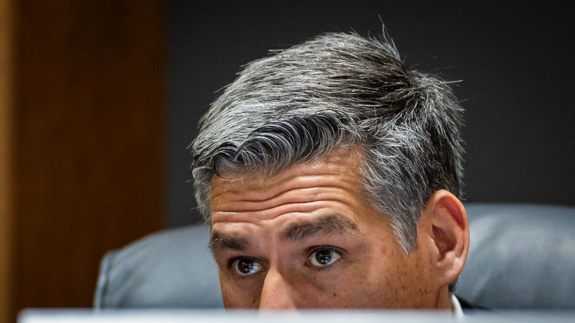 Close-up of a man with salt-and-pepper hair peeking over a desk edge, eyes looking to the left, in a dim room.