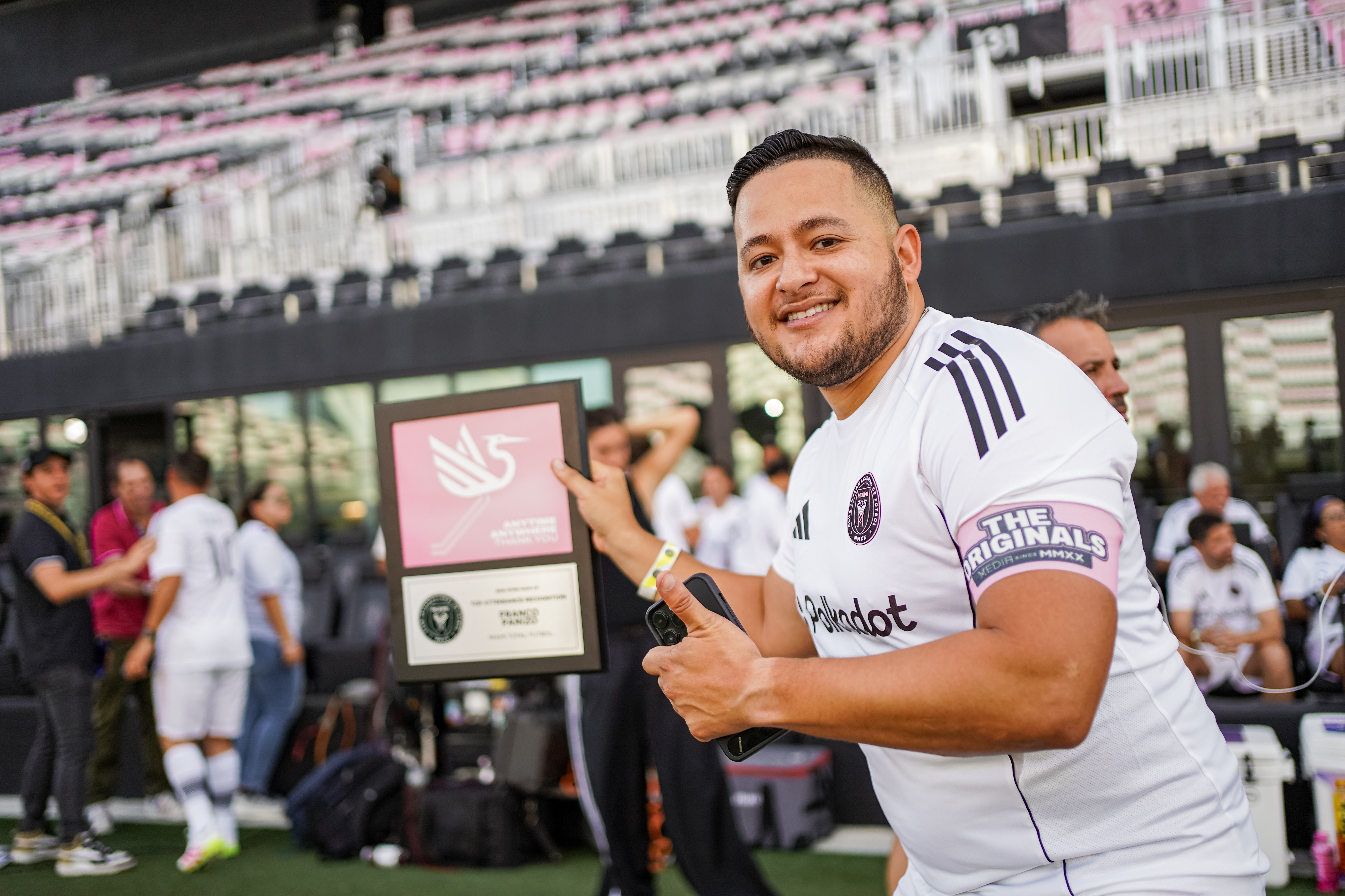Smiling man in a white sports jersey holds a framed award, standing in front of a stadium with pink seating and blurred spectators.