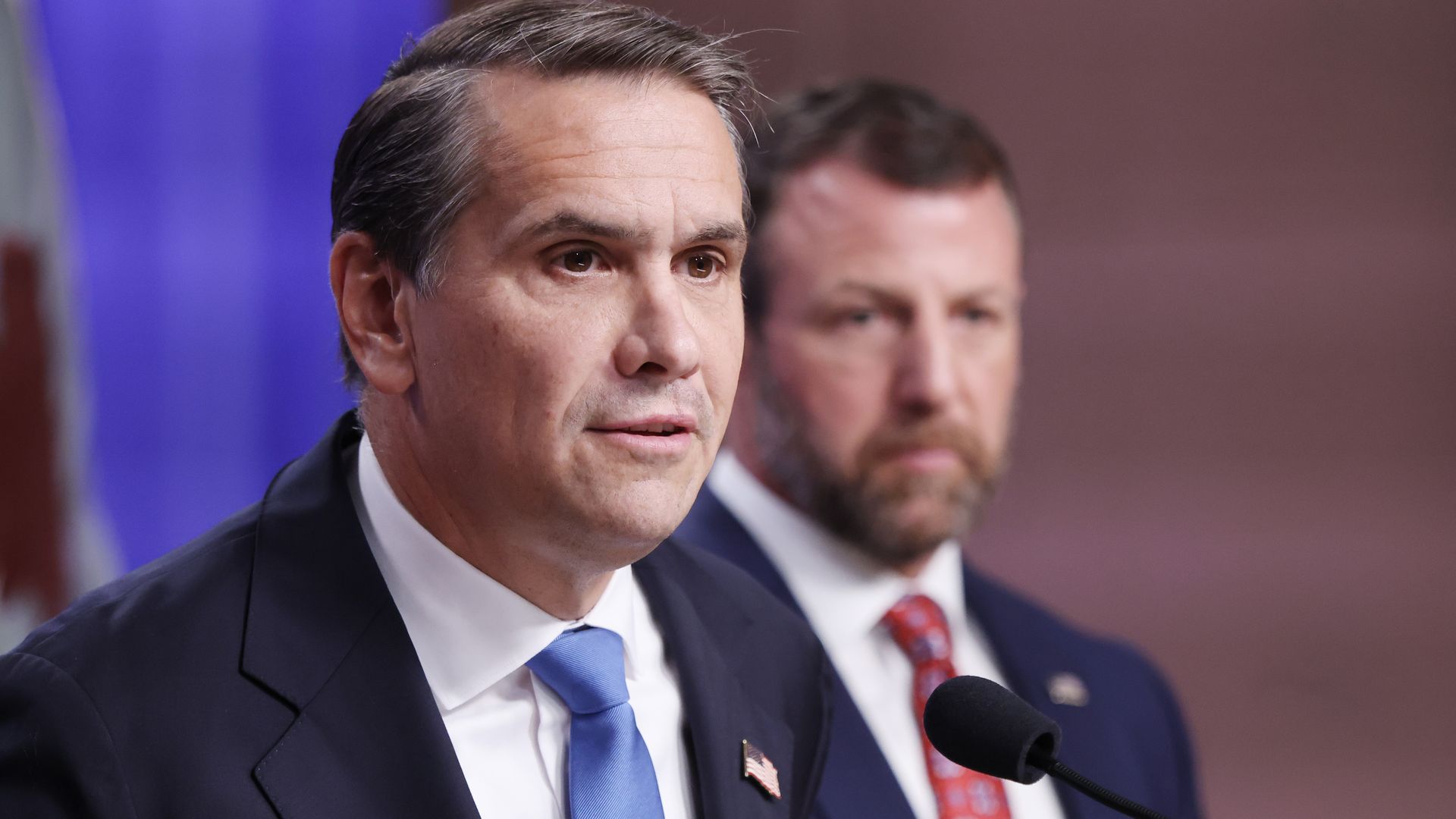 A man with dark hair wearing a dark suit, a white collared shirt, a blue tie and an American flag pin (Todd Blanche) speaks into a microphone at a news conference while another man in a blue suit, a white collared shirt and a red tie (Markwayne Mullin) stands behind.