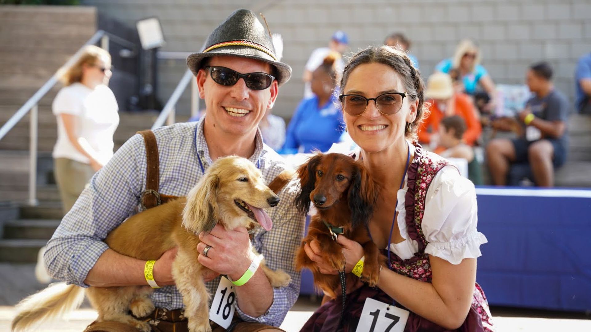 Smiling man in a gray fedora and sunglasses and smiling woman in a dirndl hold two long-haired dachshunds at an outdoor event; race numbers 18 and 17 on their outfits, with a crowd and steps behind them.