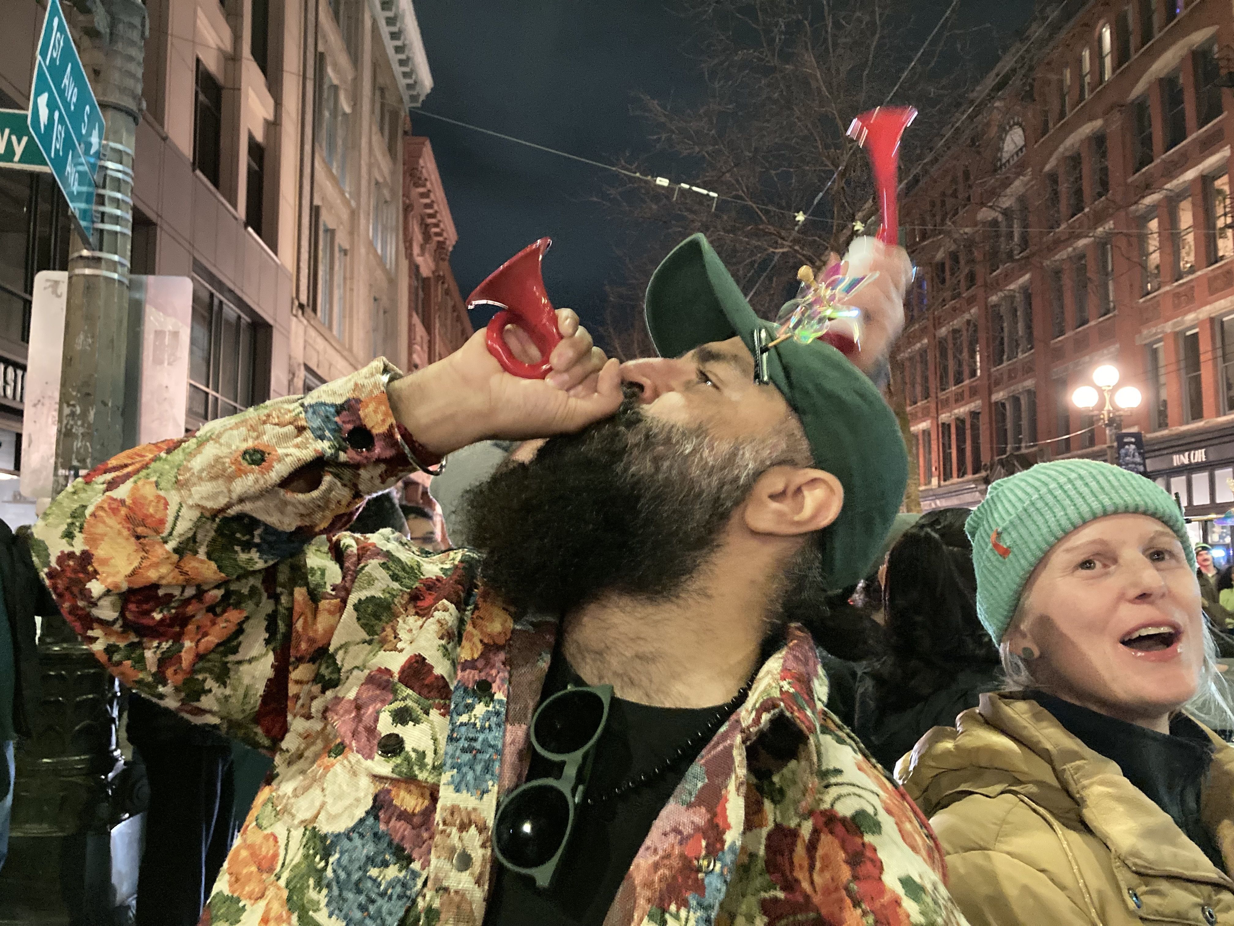 A bearded man in a floral jacket and green cap blows a red horn at a night street event, while a smiling woman in a light blue beanie and yellow jacket stands nearby in a lively city setting.