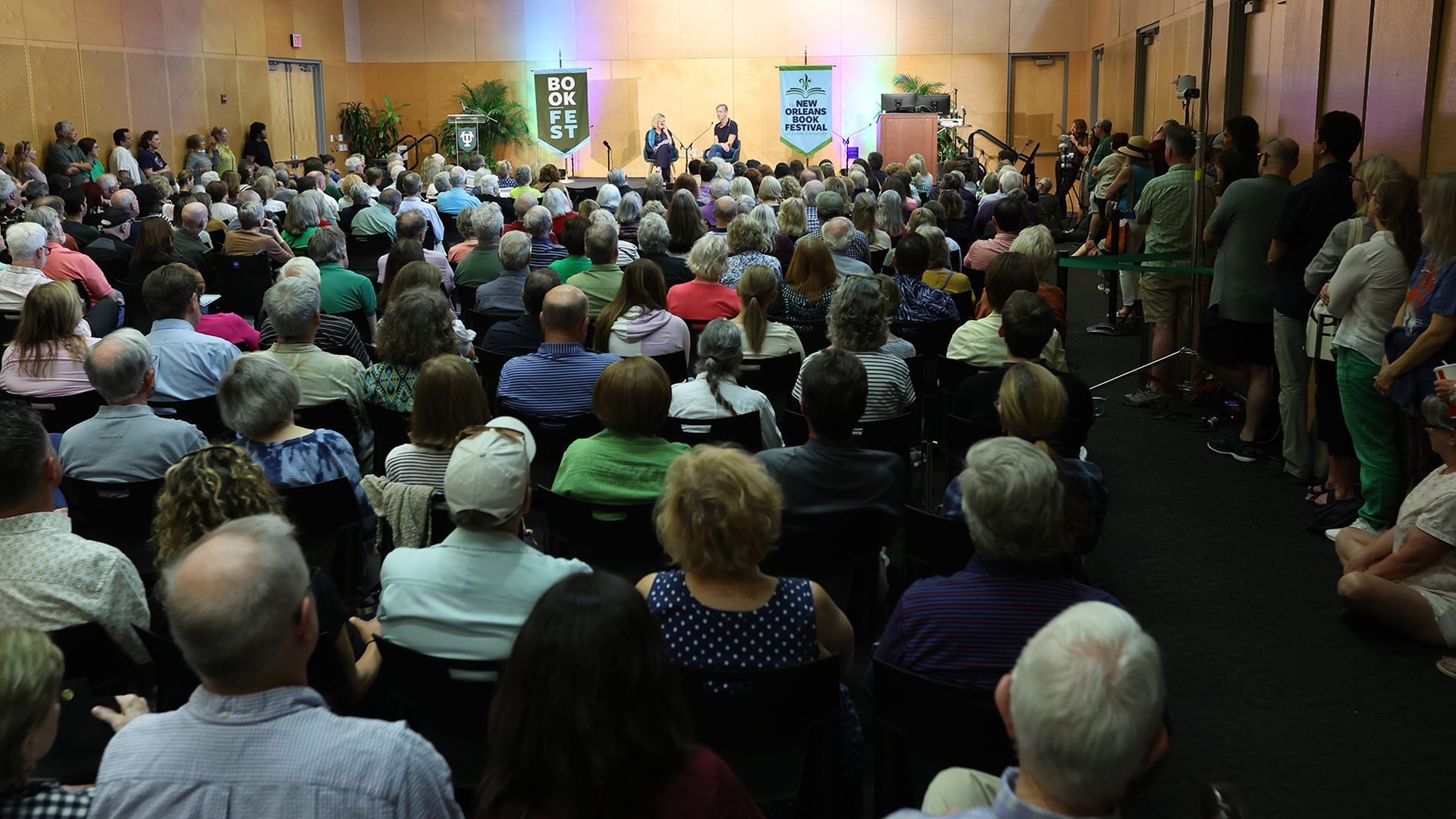 A full auditorium of people watch a conversation between two people on a stage on the far end of the photographed space.