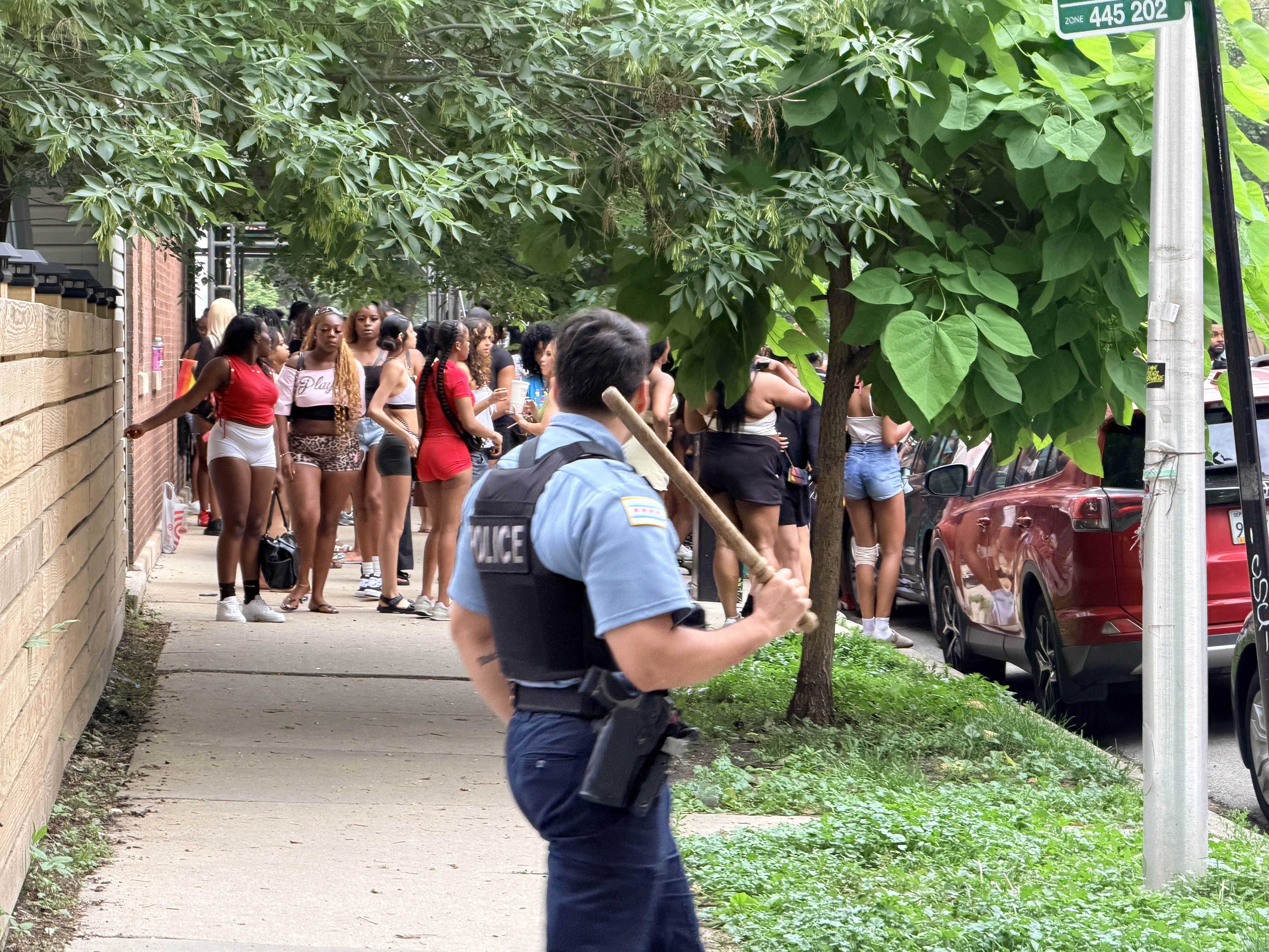 Cop with a club on a leafy block with young Black teens in the background.