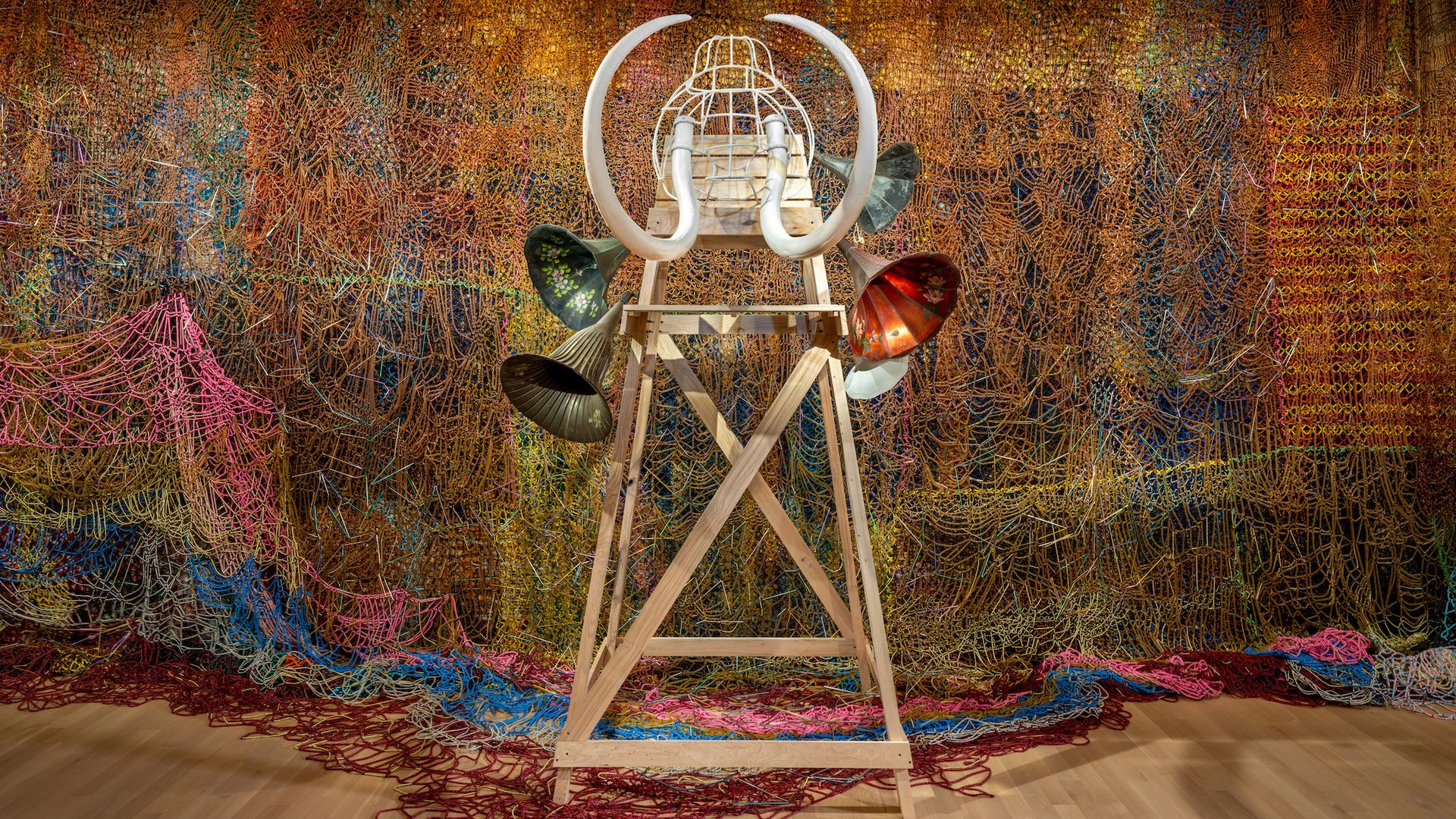Art installation featuring a tall wooden frame with a white wire cage and large curved tusks on top, surrounded by four colorful metal horn-like structures, set against a vibrant woven backdrop.