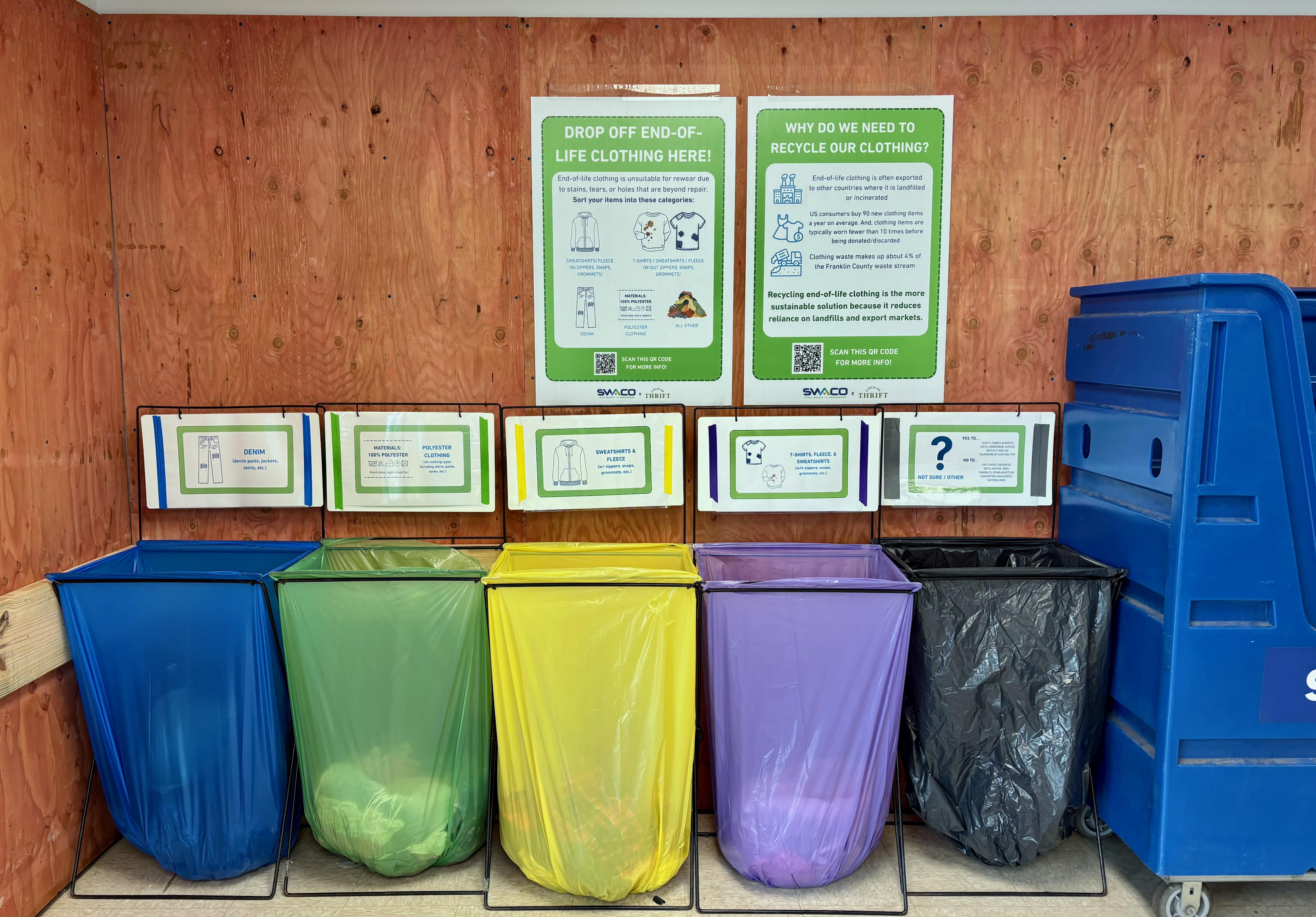 A clothing recycling station at a Columbus Goodwill store. There are five bins, each a different color, where donors can drop off clothing that has reached the end of its life. There is signage explaining how the sorting process works.