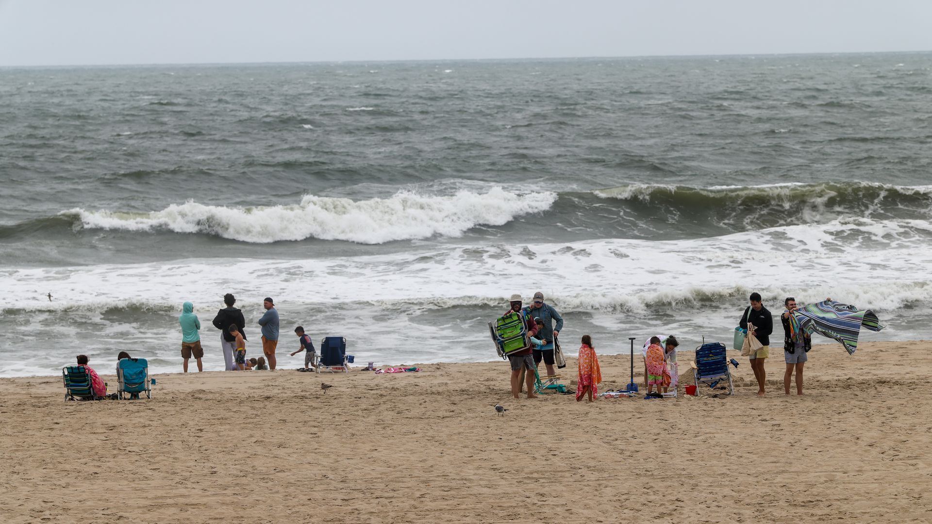 Groups of people, some wrapped in towels, on a sandy beach with rough gray ocean waves under a cloudy sky, several holding chairs or blankets.