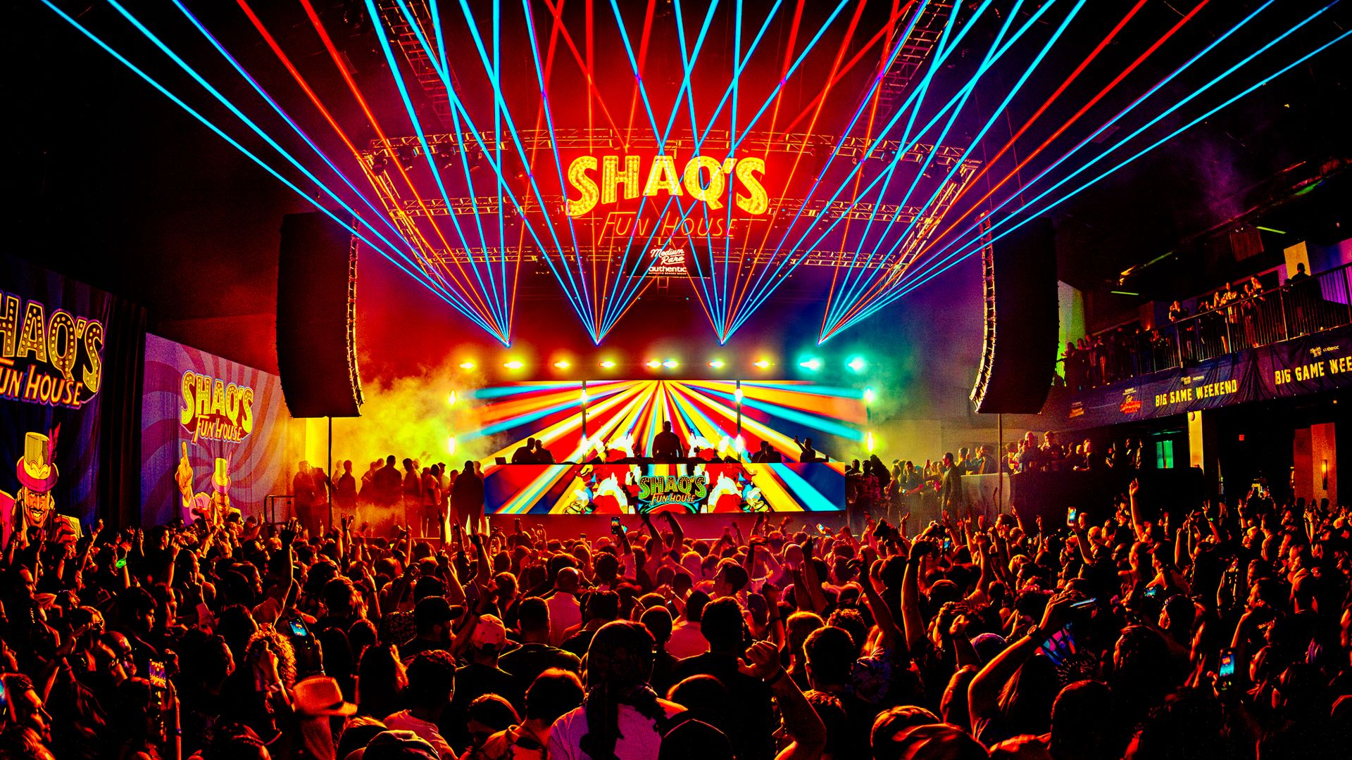 Crowded concert venue with colorful stage lights and blue laser beams, featuring large "SHAQ'S FUN HOUSE" signs above and on the DJ booth, and a cheering audience.