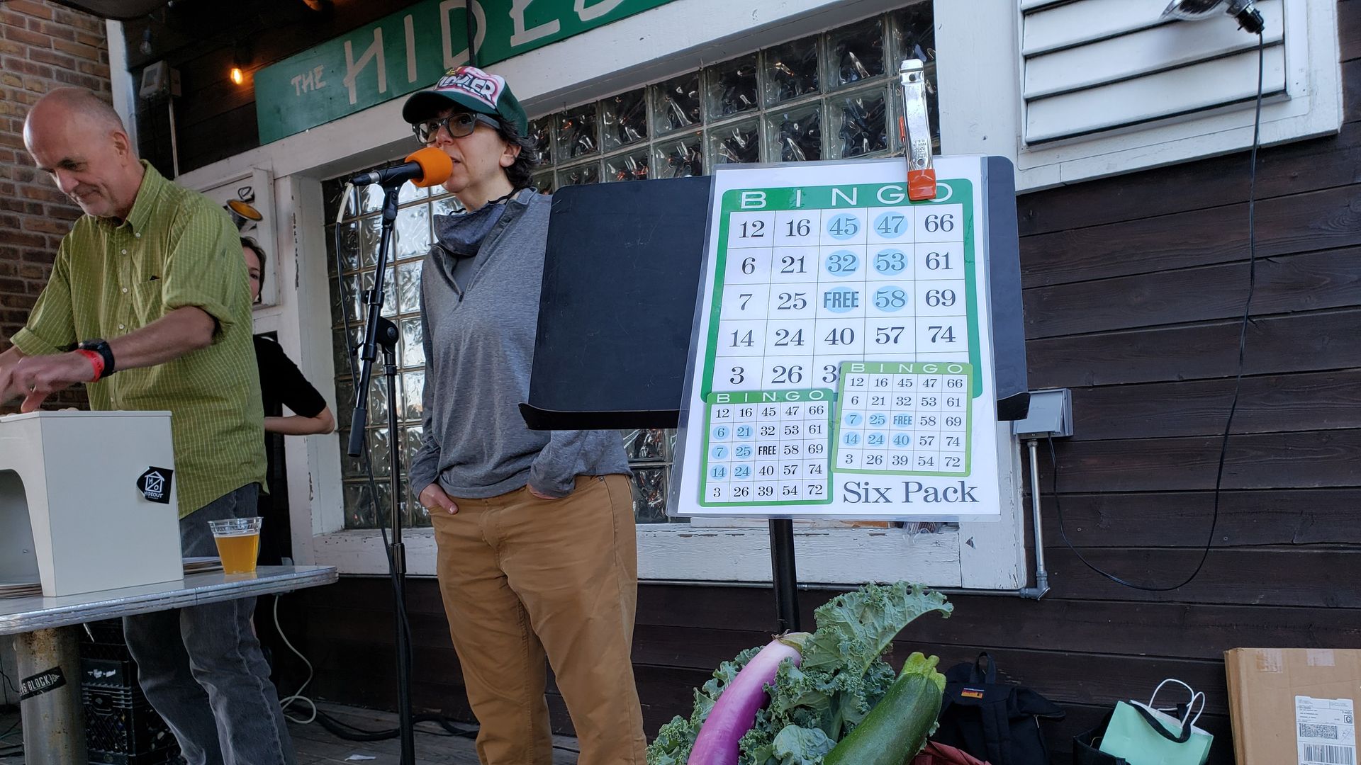 Person standing behind a microphone, next to a large BINGO card, and a basket of vegetables on table in front.